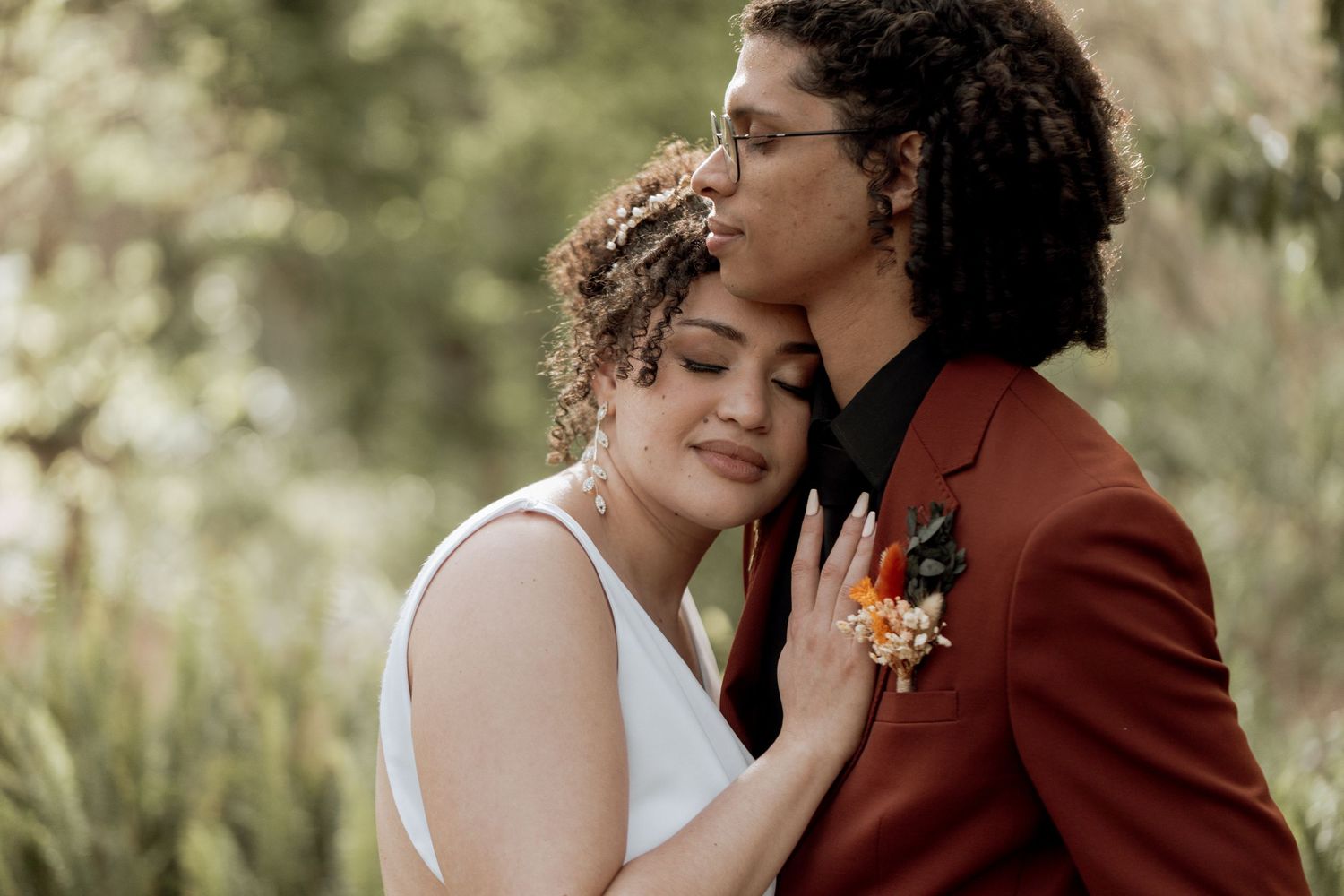 Romantic couple in wedding attire embraces lovingly in a sunlit outdoor setting with greenery in the background.