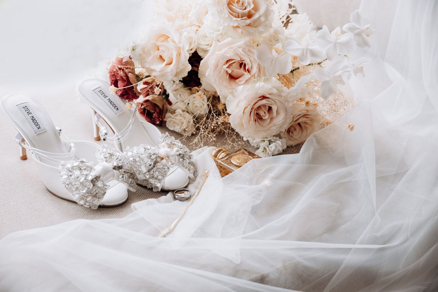 Elegant bridal flat lay featuring crystal embellished heels, a blush rose bouquet, and a flowing white veil.