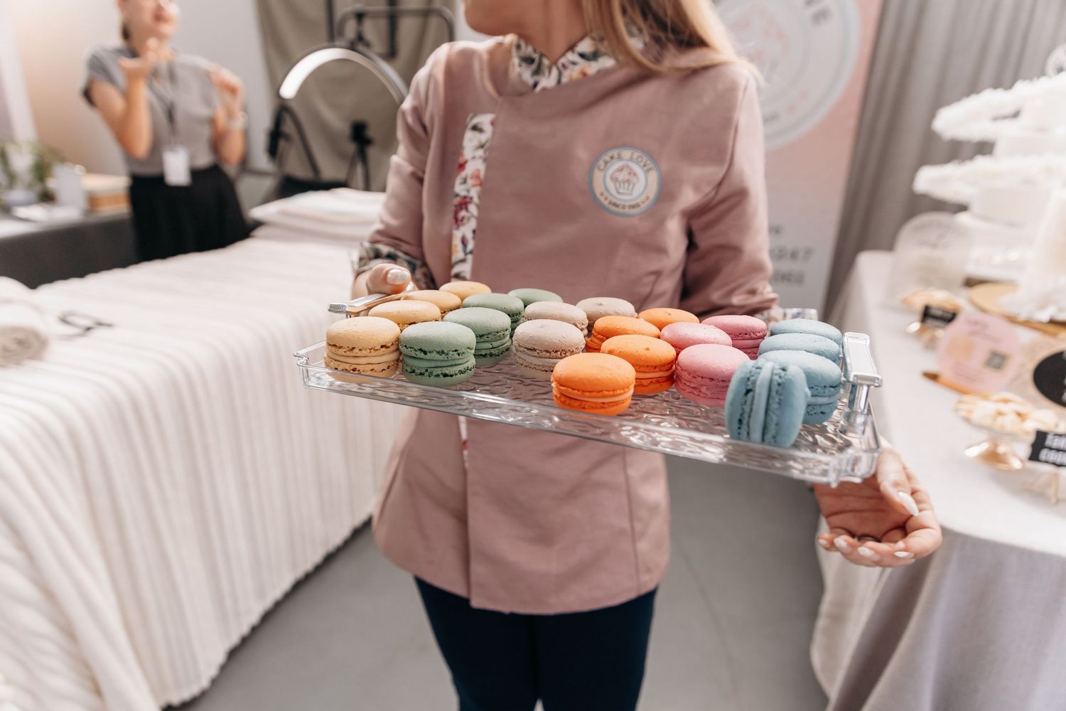 Baker in pink uniform holds tray of colorful French macarons at a dessert display event.