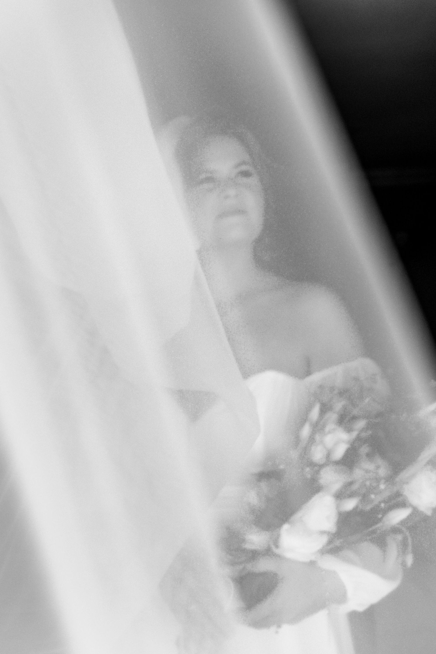 Black and white bridal portrait of a bride holding a bouquet, partially obscured by a flowing white veil.