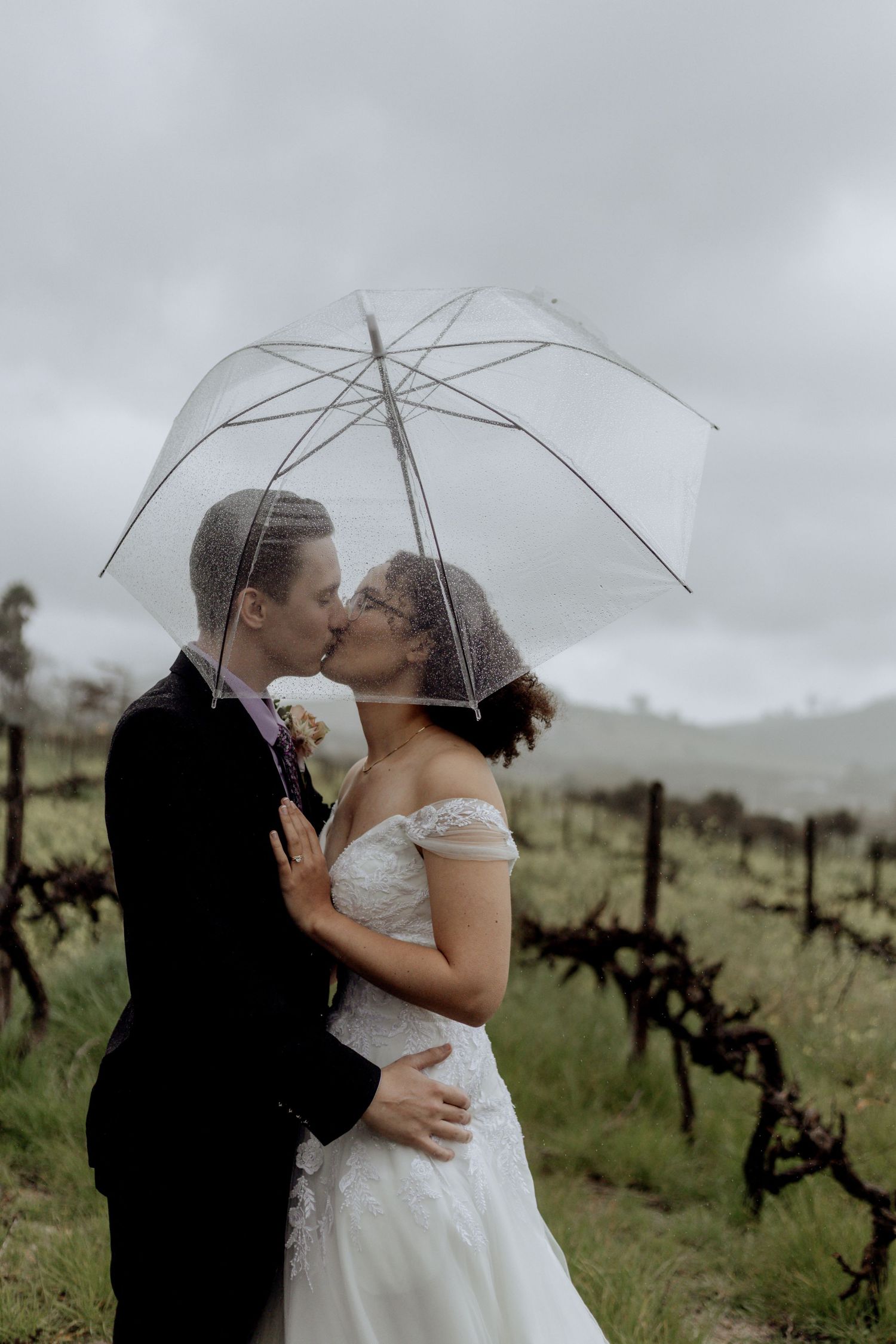 Romantic couple shares kisses under umbrellas in a dreamy vineyard setting on their rainy wedding day.