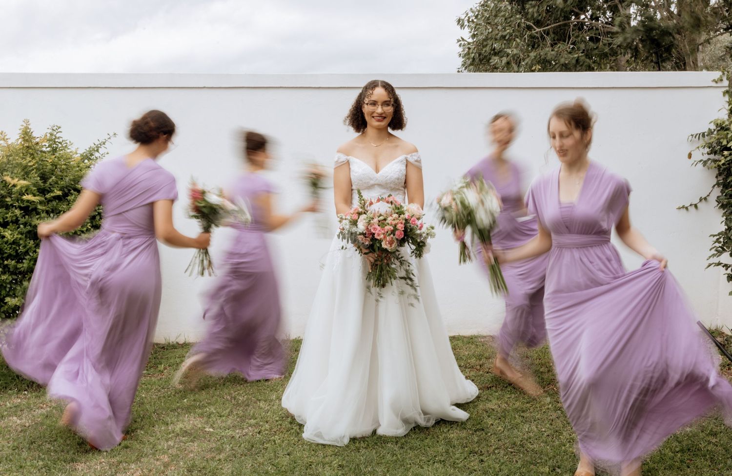 Elegant bridal party in lavender purple dresses playfully twirls and dances together on a grassy lawn.