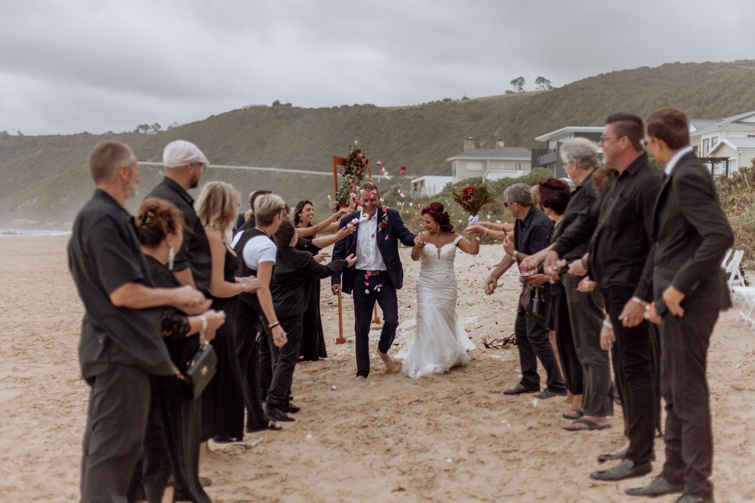 A wedding celebration takes place on an overcast beach with people lined up along both sides throwing flower petals.
