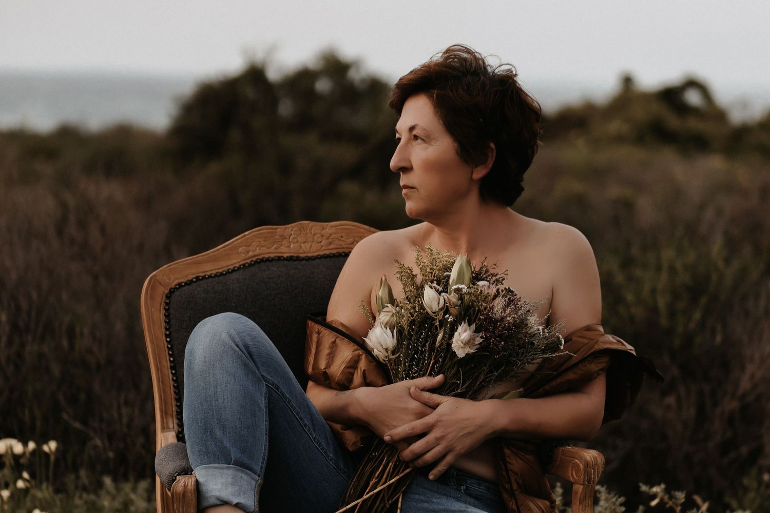 A person sits on a vintage brown leather chair in a moody outdoor setting with dried grass and cloudy skies.