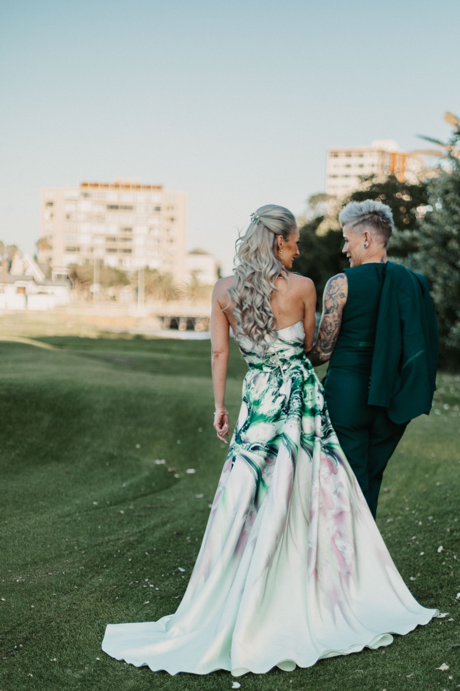 Couple in formal attire poses together on a green lawn with buildings visible in the background.