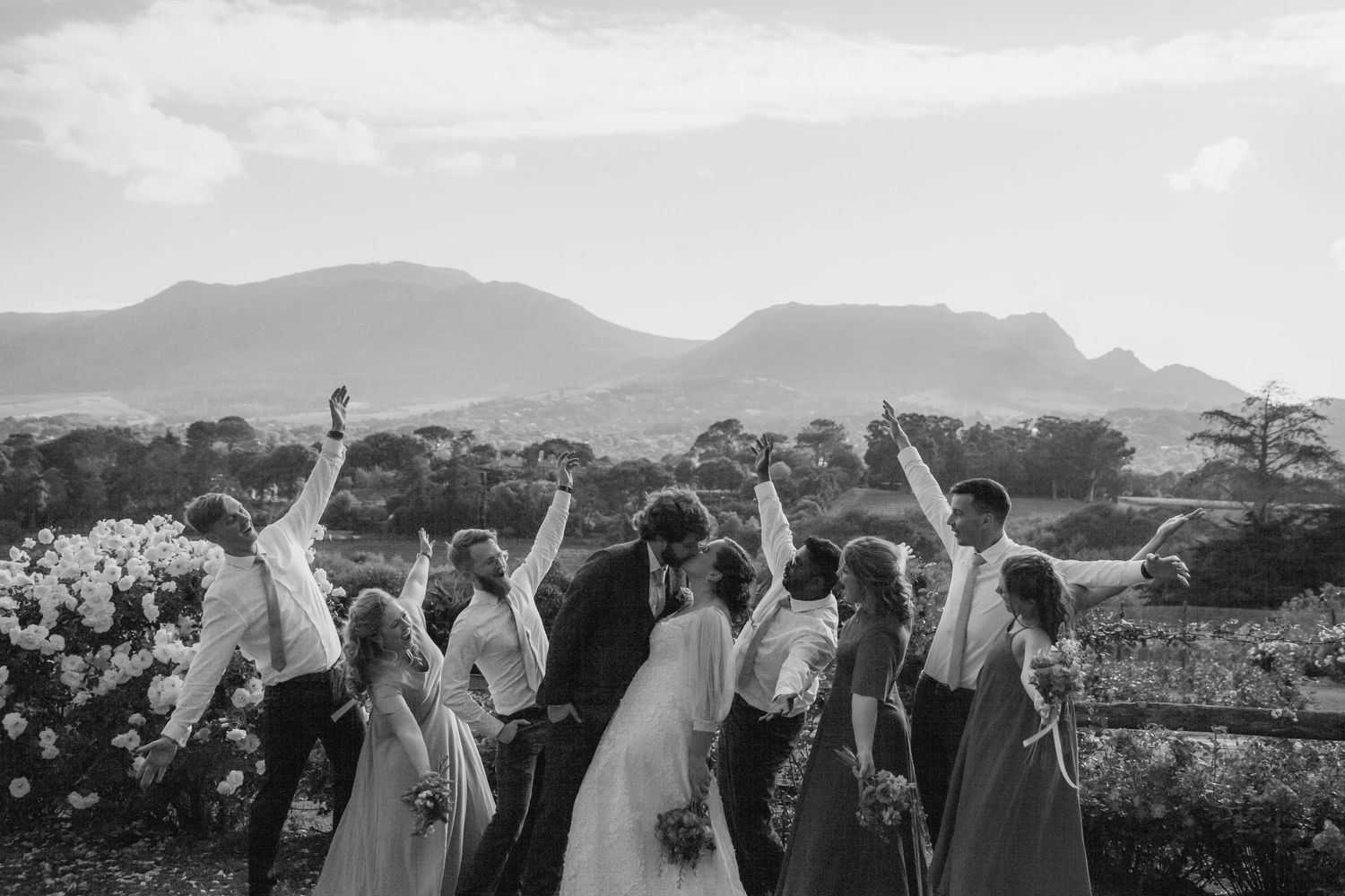 Wedding guests raise their arms in celebration against a scenic mountain backdrop at an outdoor ceremony.