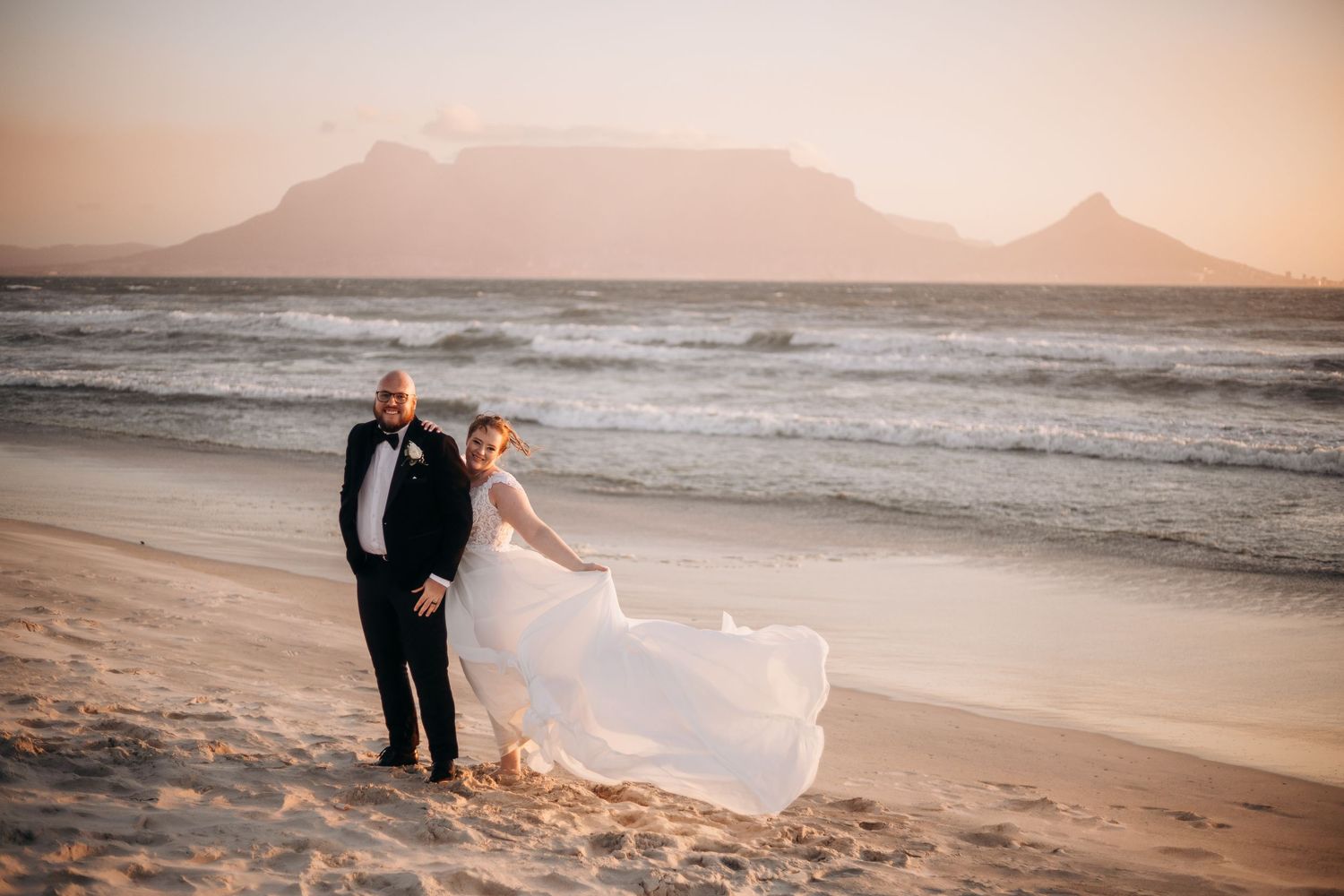 Bride and groom walk on sandy beach at sunset with Table Mountain backdrop in Cape Town, South Africa.