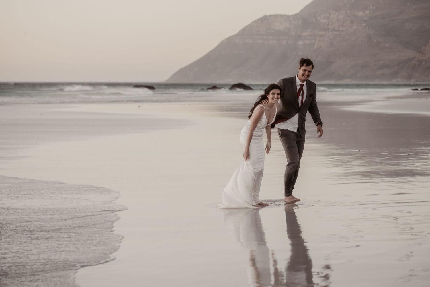 Newlywed couple walking hand in hand on a reflective beach at sunset with mountains in the background.