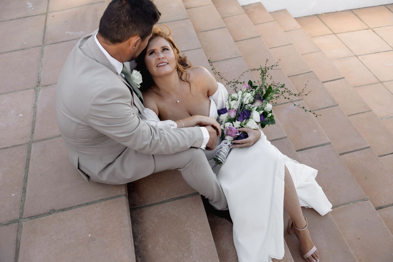 Bride and groom sitting on stone steps, bride holding a purple and white bouquet in a white gown.