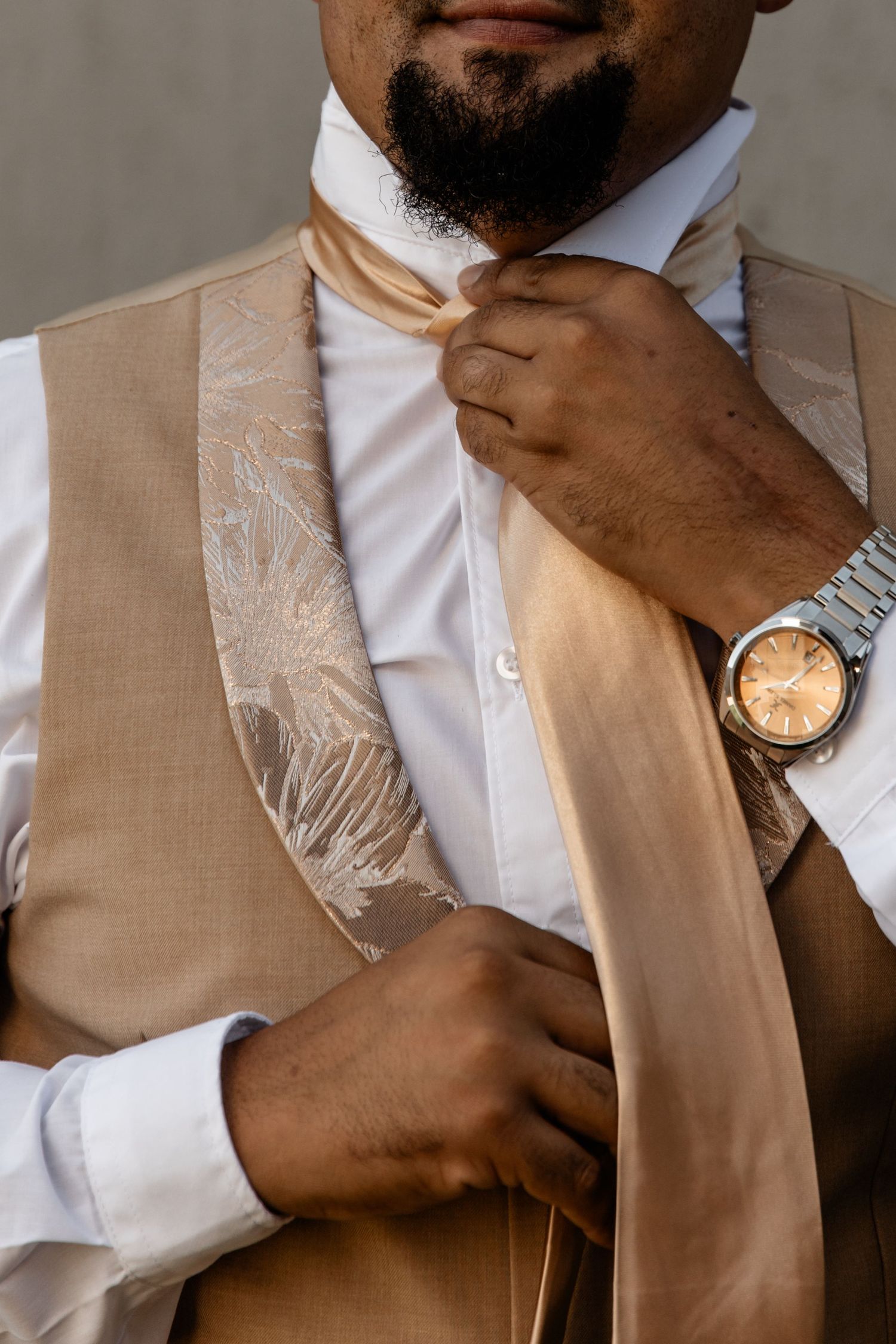 A person adjusts their beige tie while wearing a matching vest, white shirt, and silver watch on their wrist.