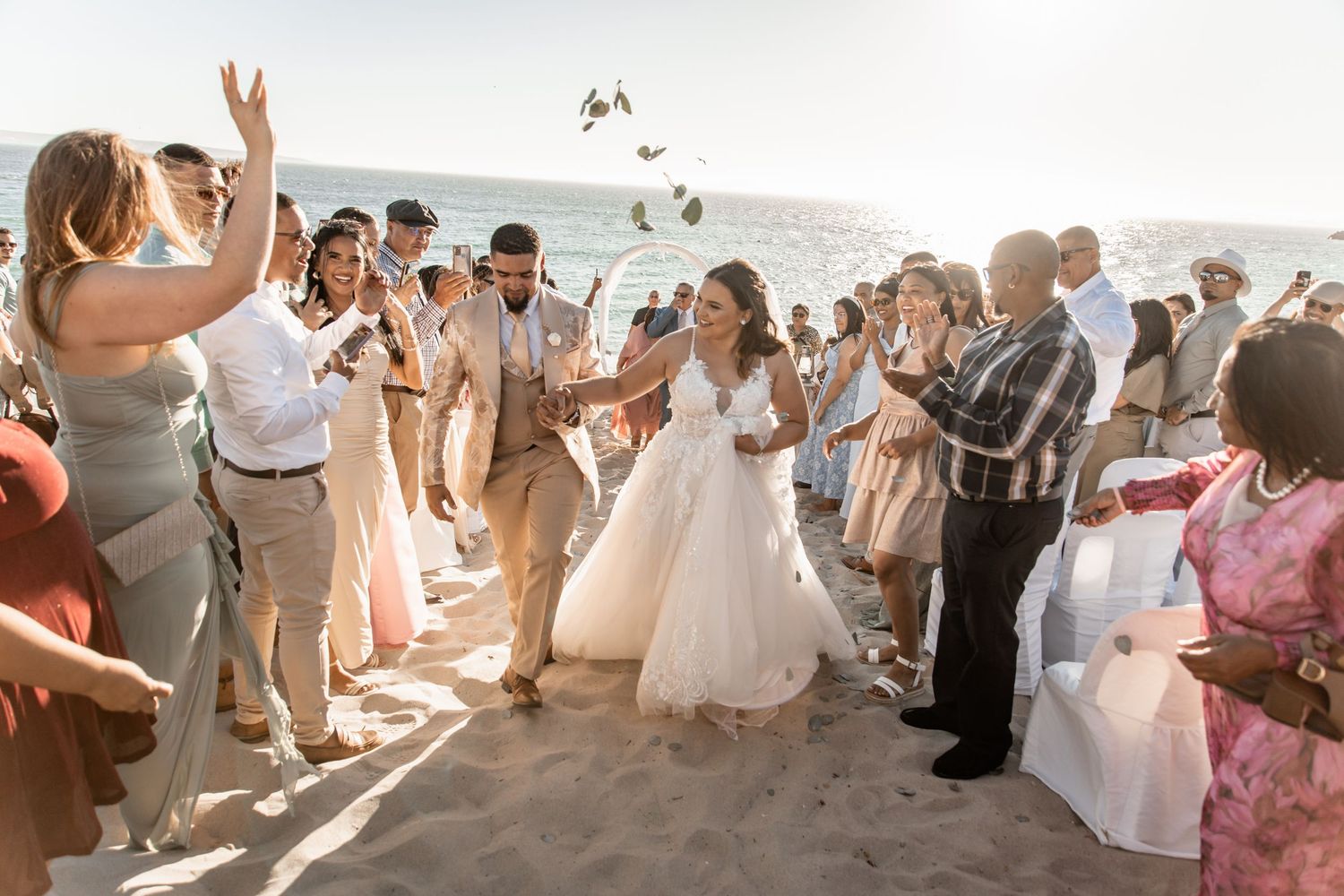 Newlyweds walk through crowd of cheering guests tossing flower petals during sunset beach wedding celebration.