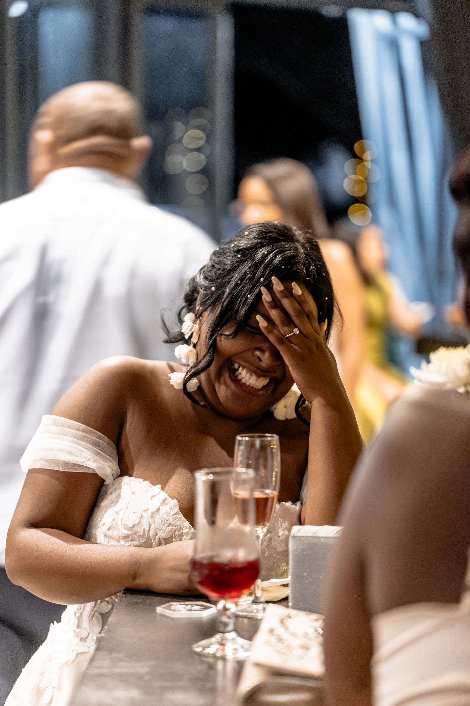 A person laughs joyfully at a social gathering while seated at a restaurant table with drinks visible.