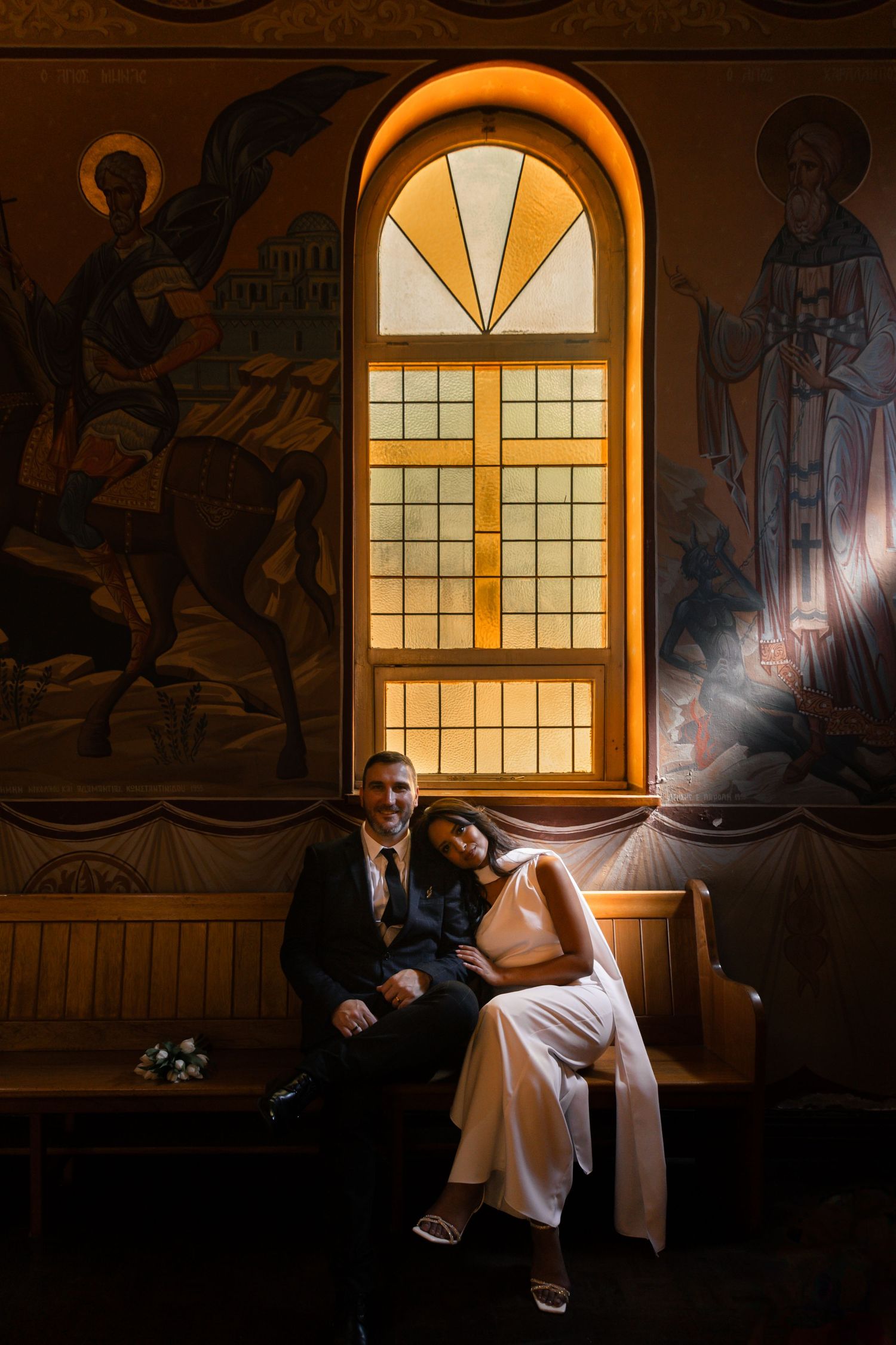 Bride and groom sitting together on a wooden church pew beneath a glowing arched stained glass window with religious murals.