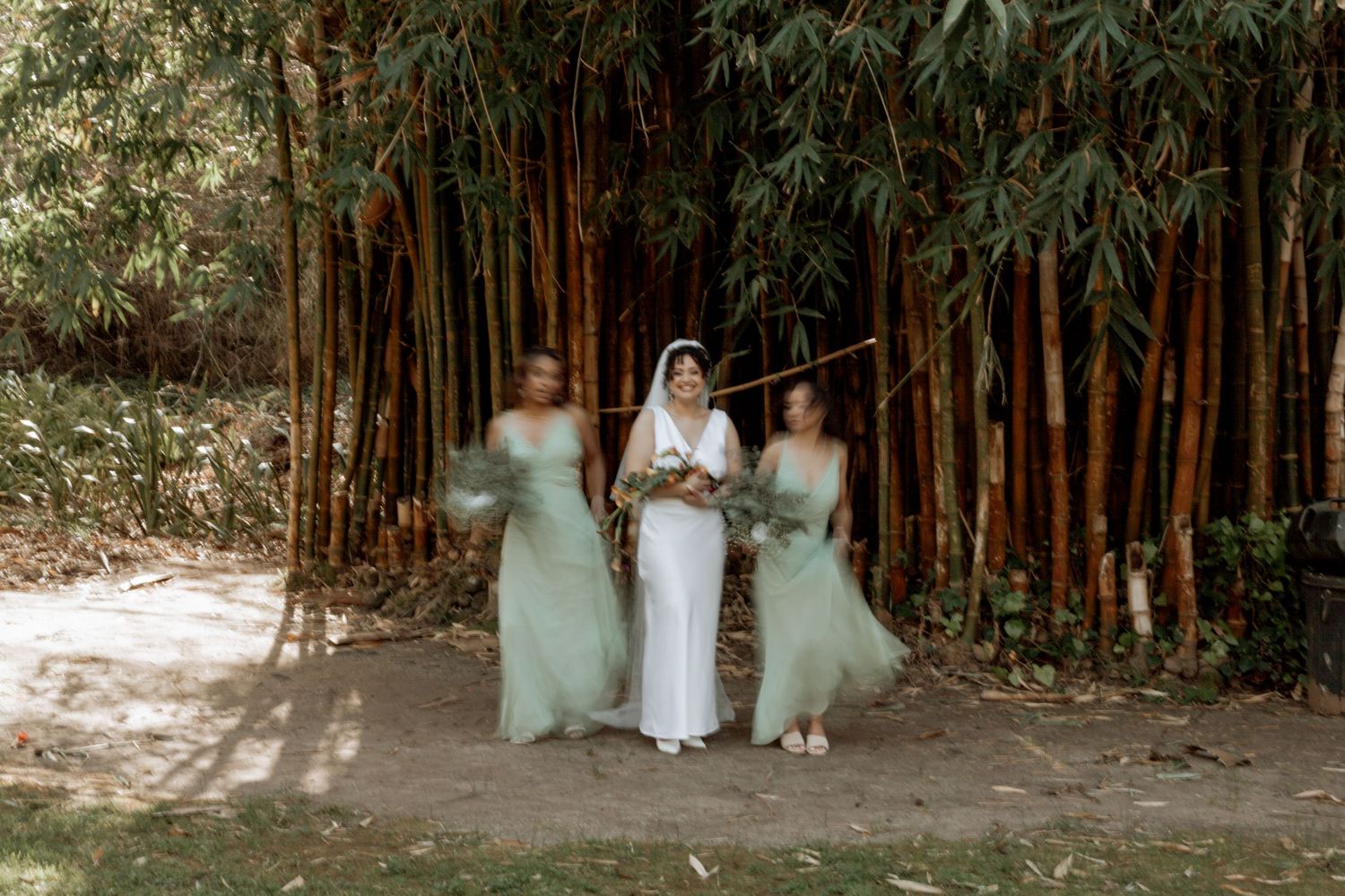 Elegant wedding party wearing white and mint green dresses pose in front of a bamboo grove on a sunlit path.