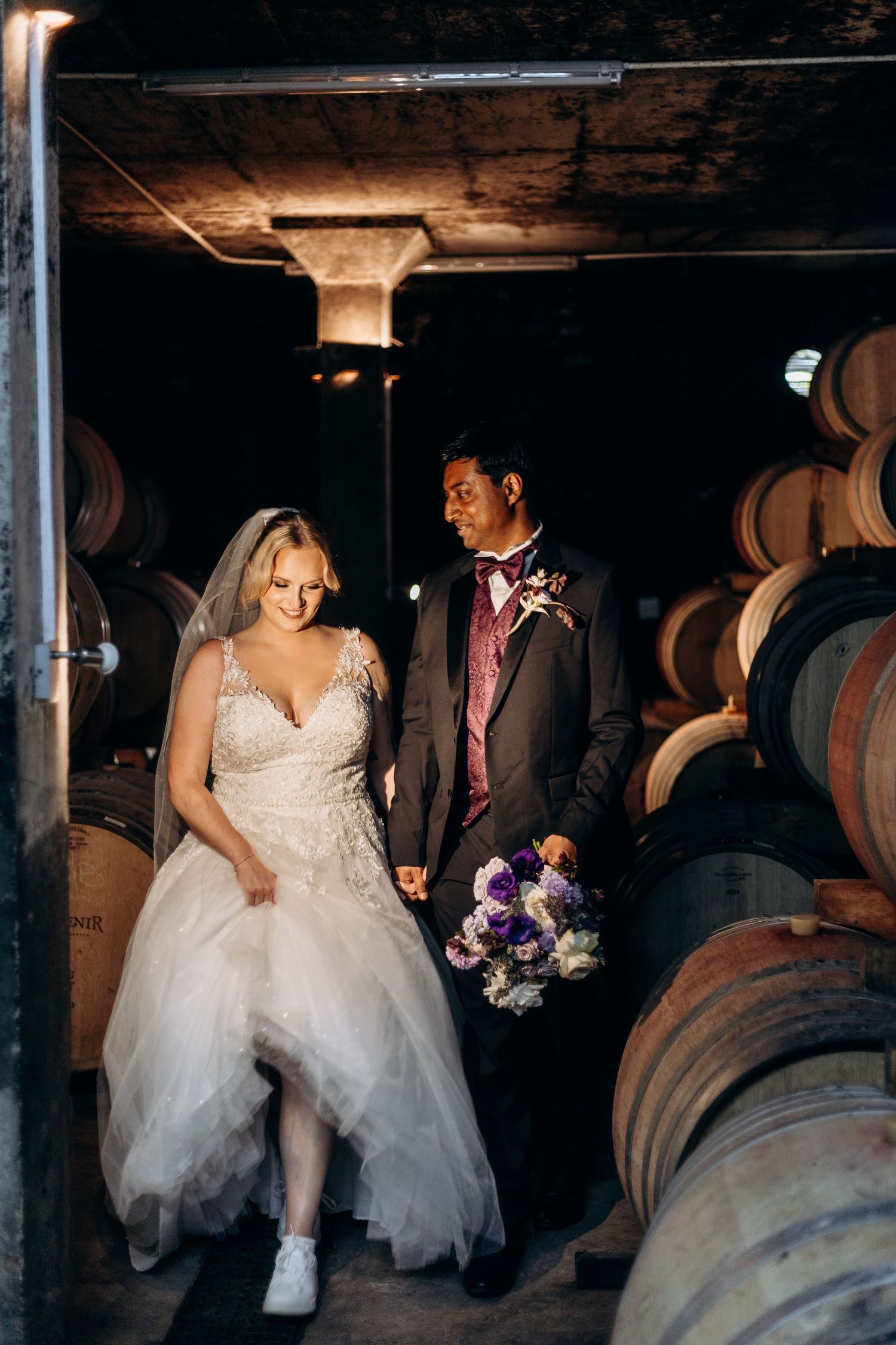 Bride and groom walk between wine barrels in a dimly lit winery cellar, bride in tulle gown holding purple bouquet.