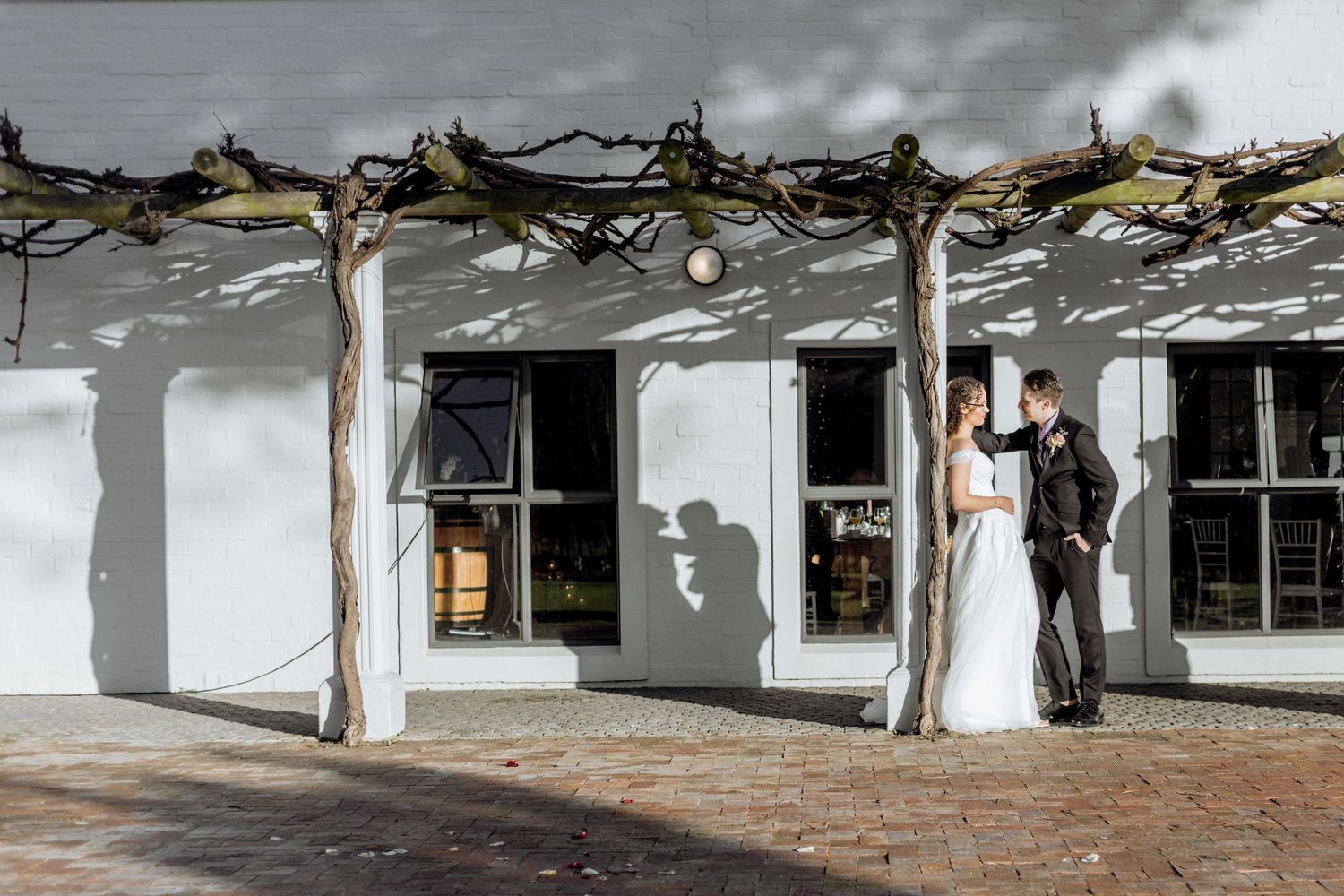 A couple poses elegantly under a rustic white porch with wooden beams and dramatic shadows at a charming venue.