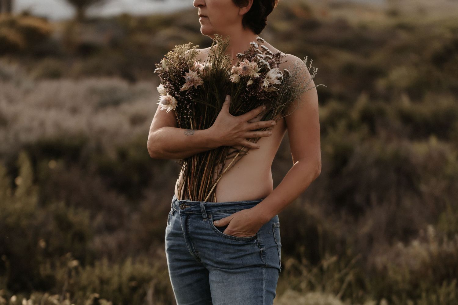 Someone holding dried florals and grass while wearing blue jeans in a moody sunset field setting.