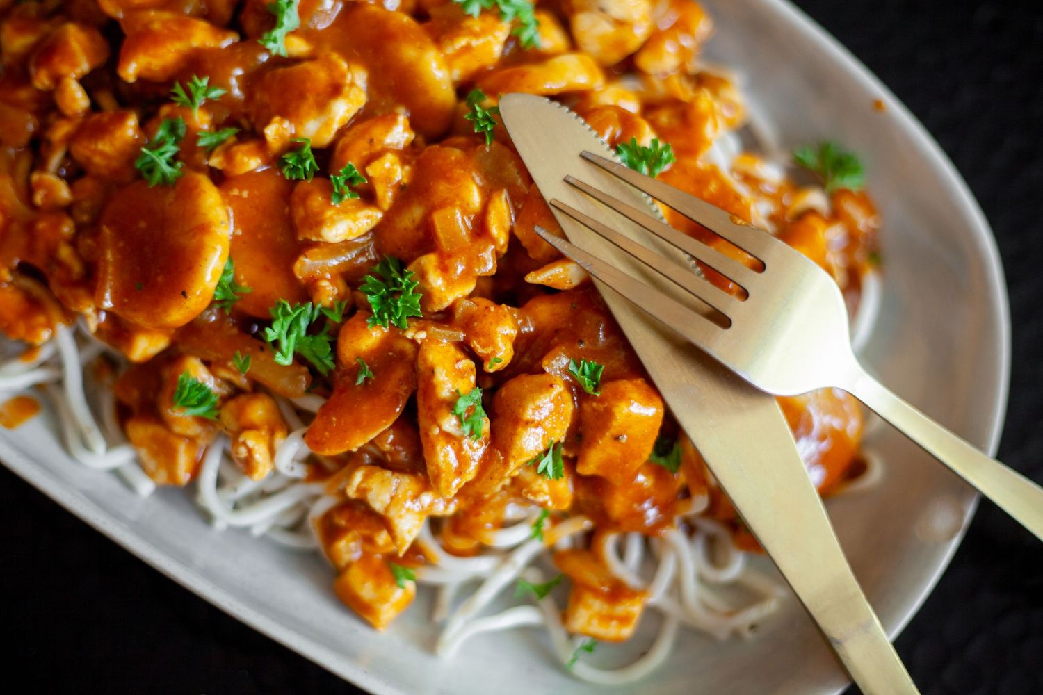Spicy orange chicken served over rice noodles garnished with fresh herbs and served with wooden utensils.
