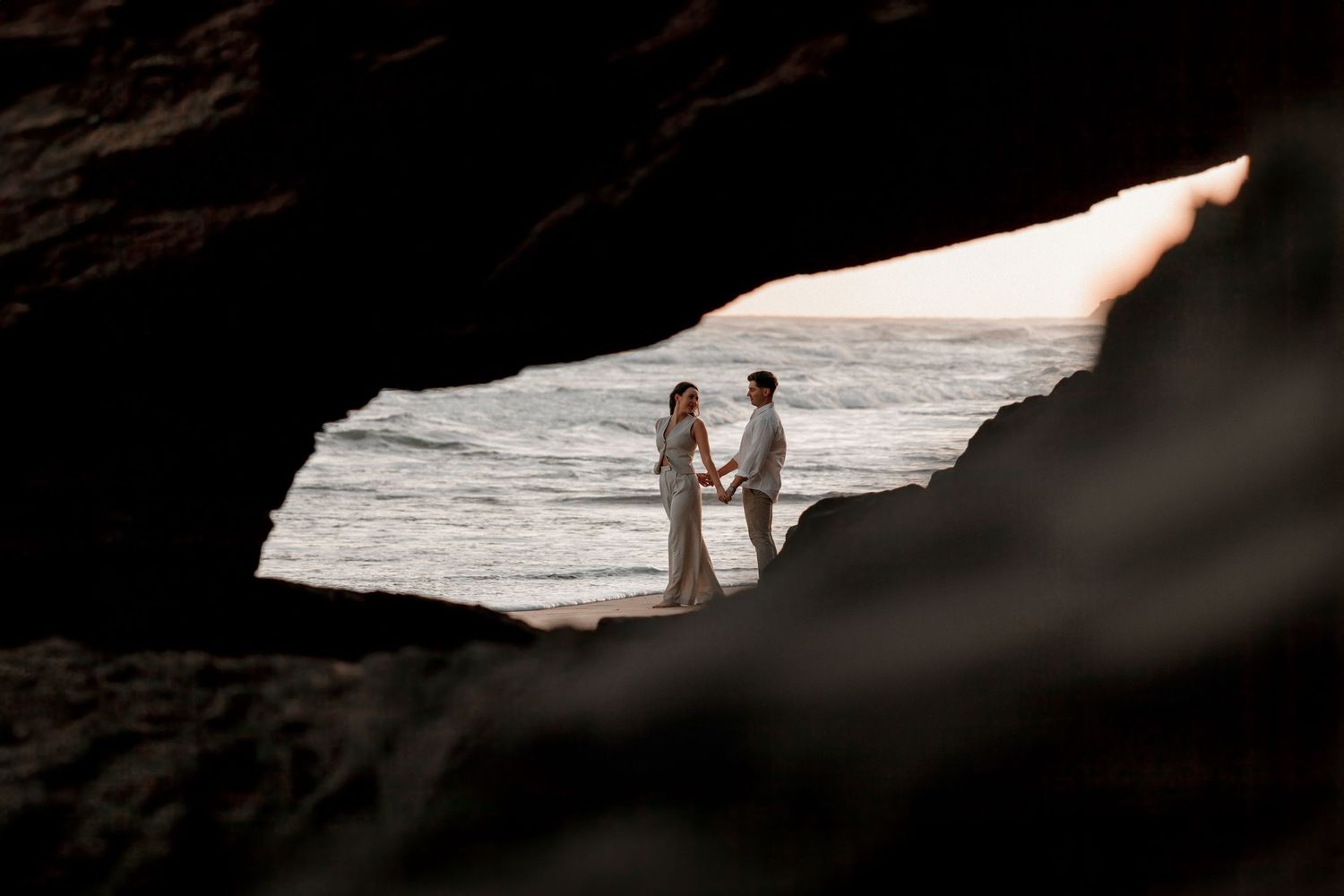 Two silhouetted figures stand together in a rocky cave opening overlooking a bright ocean view.