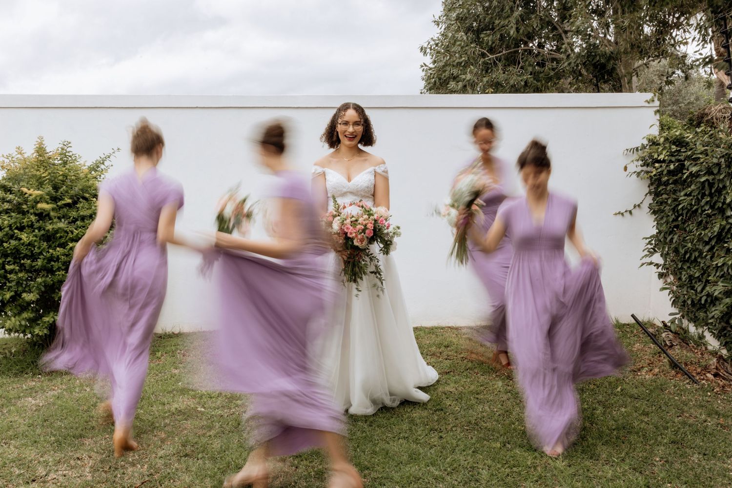 Bridesmaids in flowing lavender dresses twirl and dance on a grassy lawn during a wedding celebration.
