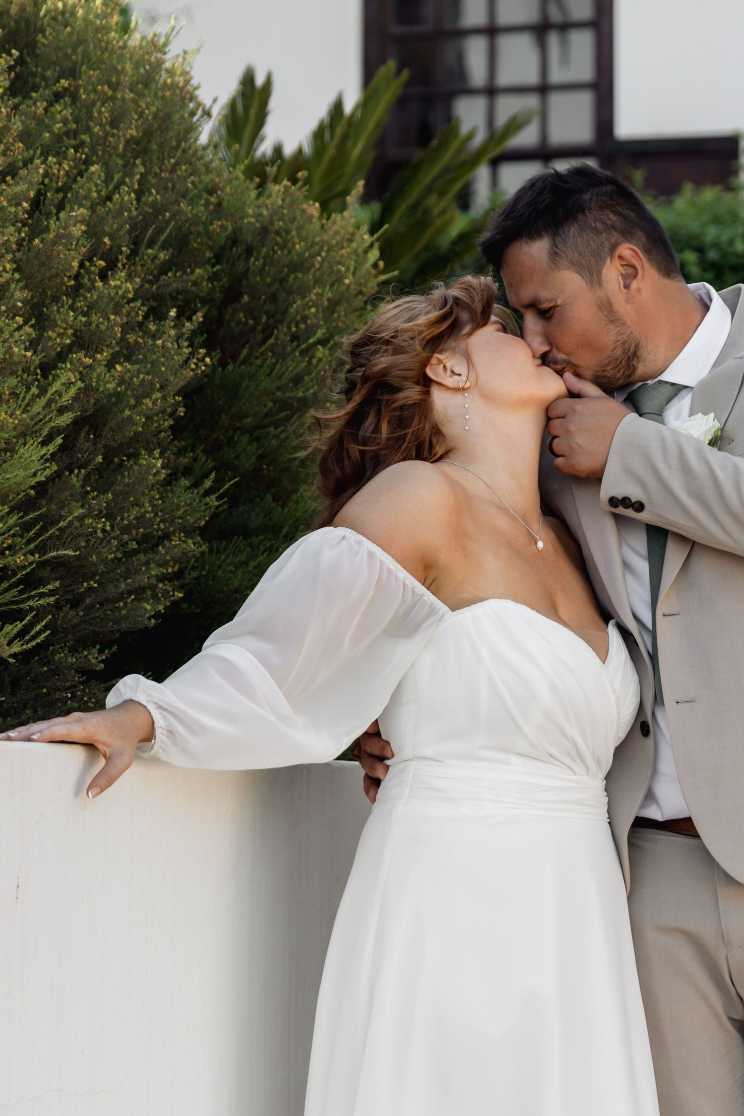 A bride in a white off-shoulder gown and groom in a tan suit share a romantic kiss outdoors near lush greenery.