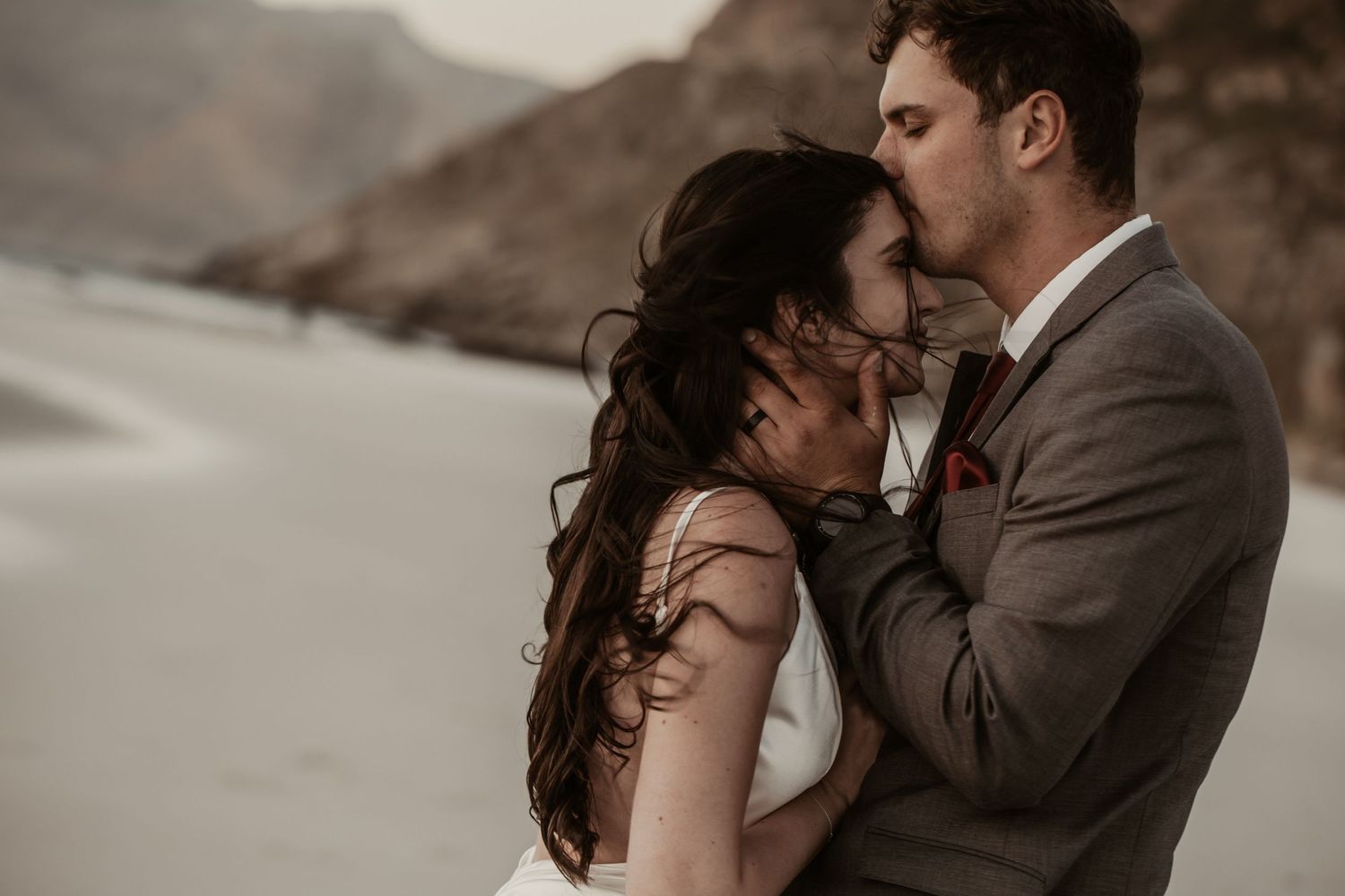 A bride and groom share an intimate moment on a beach, with the groom tenderly holding her face as they nearly kiss.