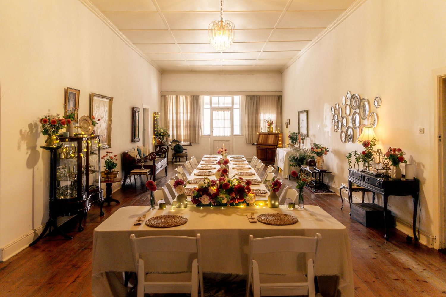 Elegant dining room with long table set for a formal dinner, floral centerpieces, white chairs, and warm lighting.