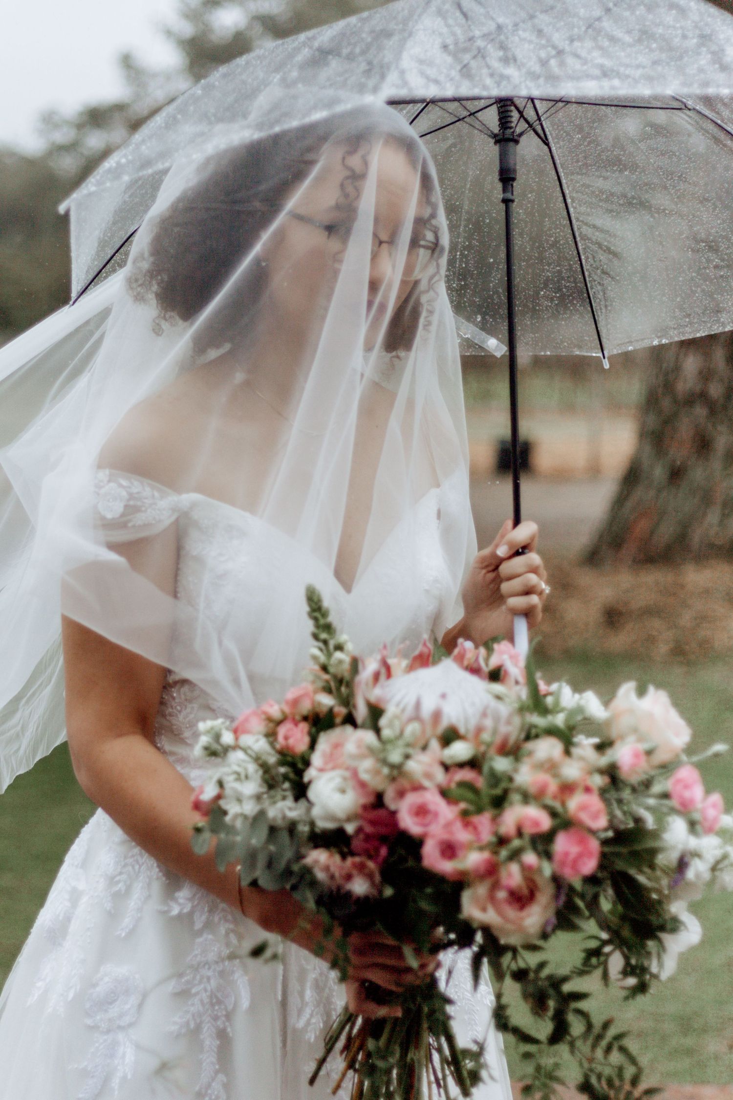 A bride holds a clear umbrella and pink floral bouquet while wearing a delicate veil on a rainy wedding day.