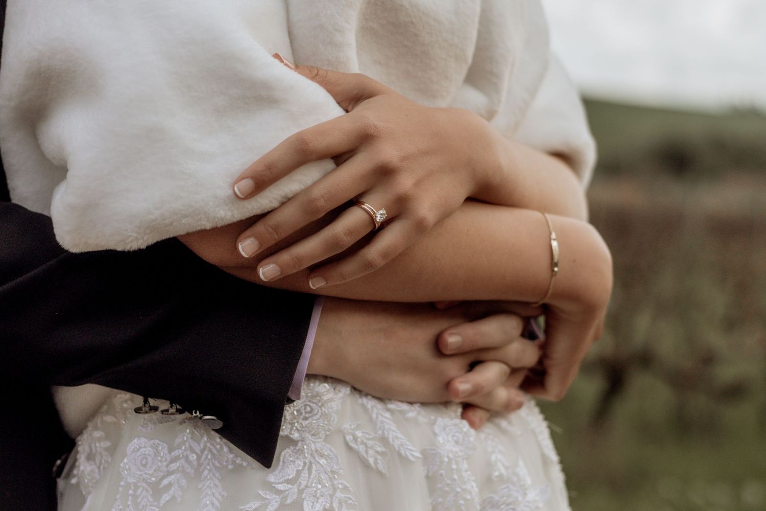 White floral wedding dress skirts swaying in a gentle breeze with ring-adorned hands clasped together.
