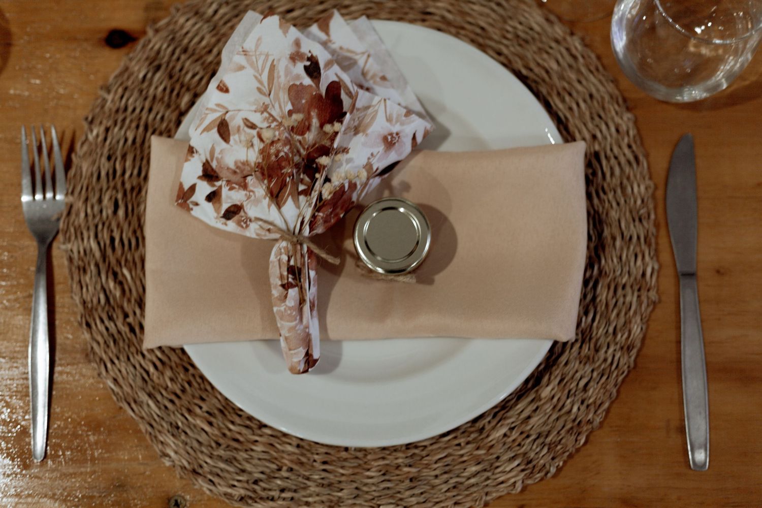 Rustic table setting with white plate on woven placemat, featuring floral napkin decor and minimalist silverware.