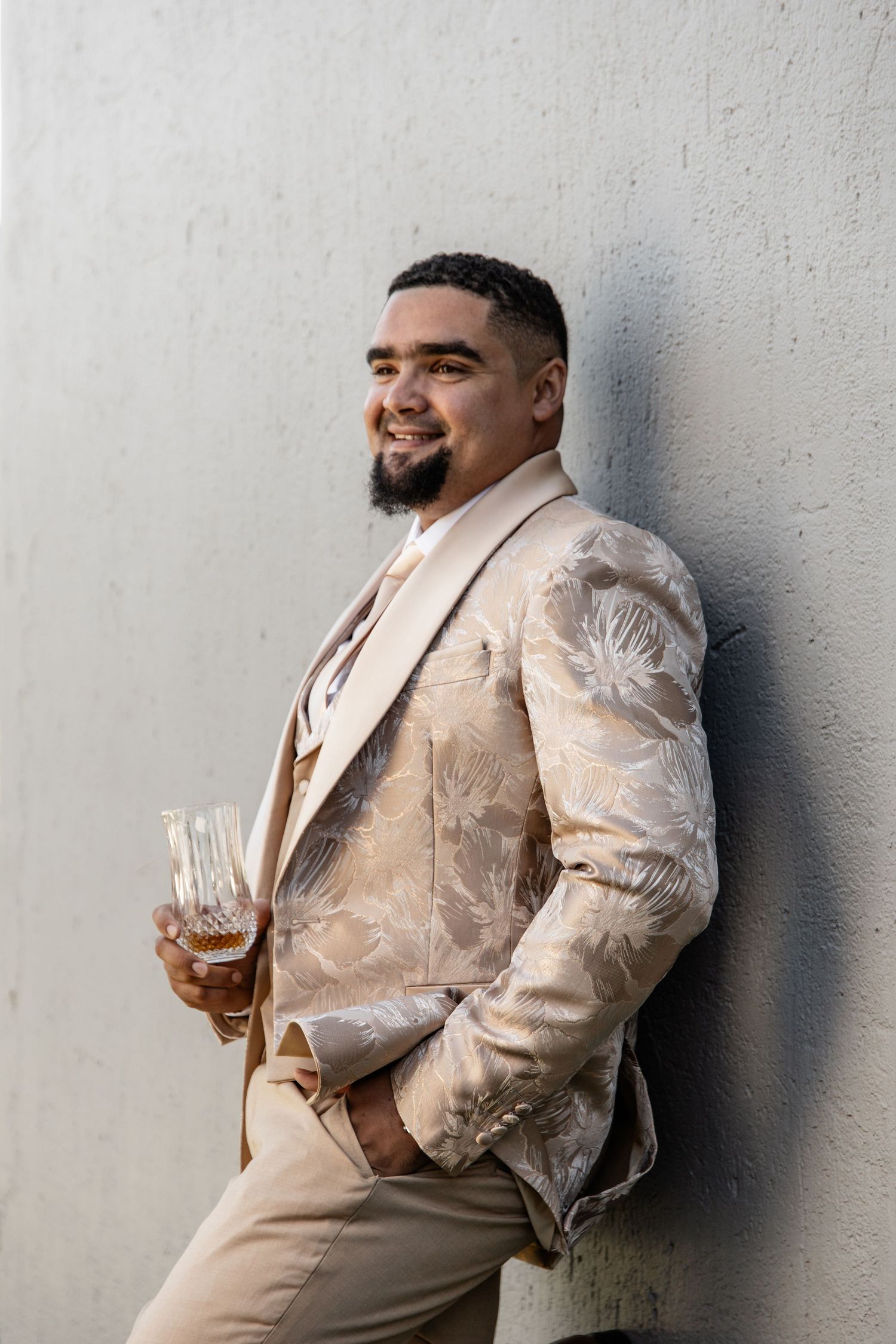 A figure in a cream-colored suit leans against a light grey wall while holding a glass of liquid.