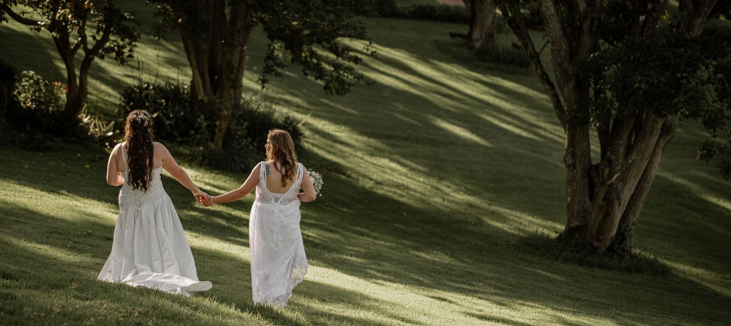 Two people in flowing white dresses walk together on a sunlit grassy hillside surrounded by trees.