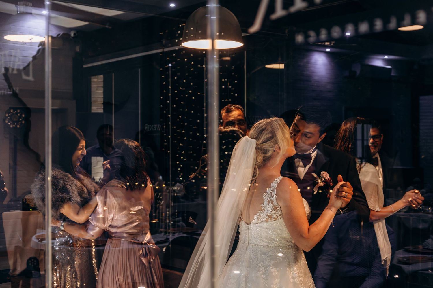 Bride and groom share first dance surrounded by guests at elegant indoor wedding reception with sparkling lights.