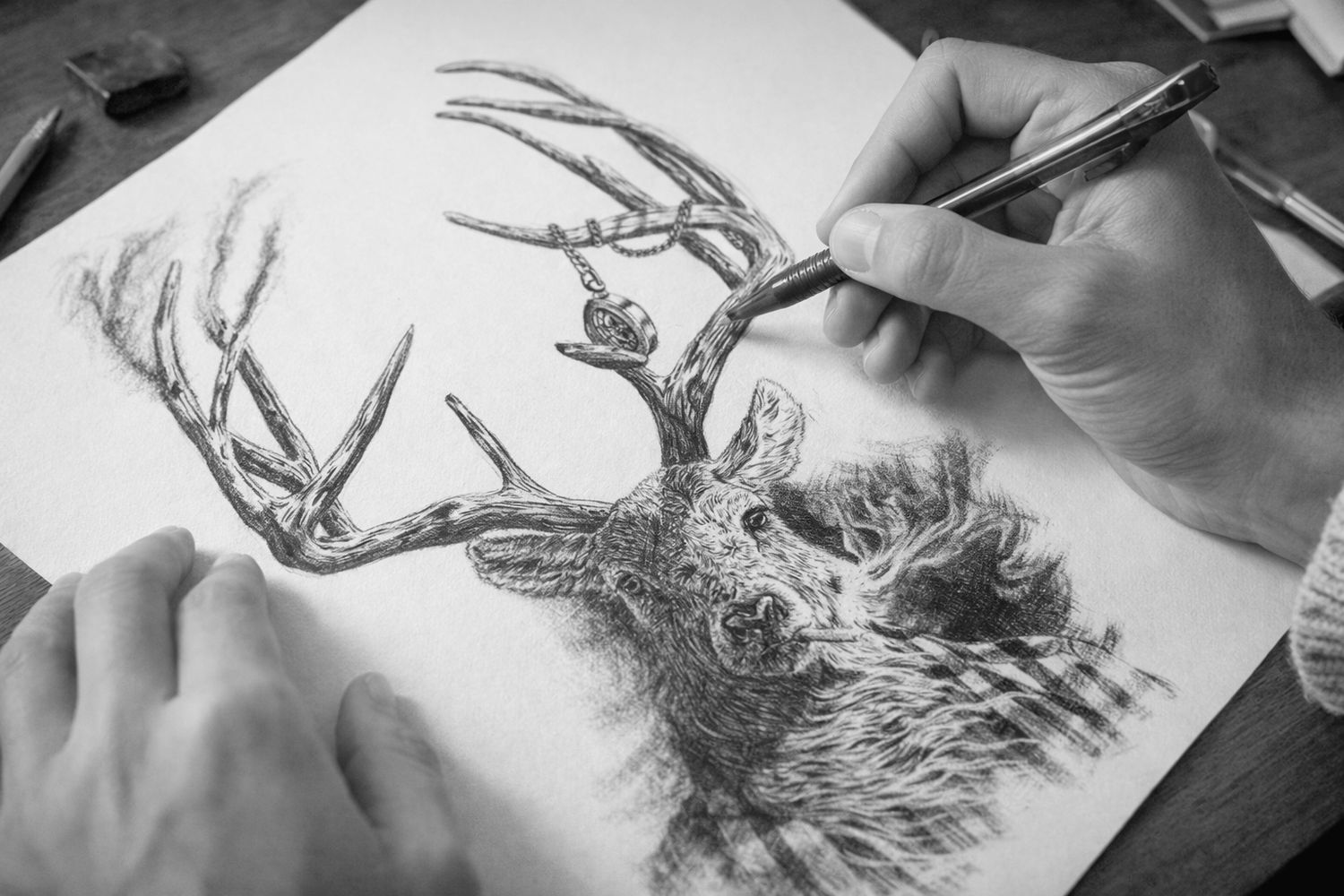 Black and white photo showing hands drawing a detailed deer with large antlers on paper using a pen or pencil.
