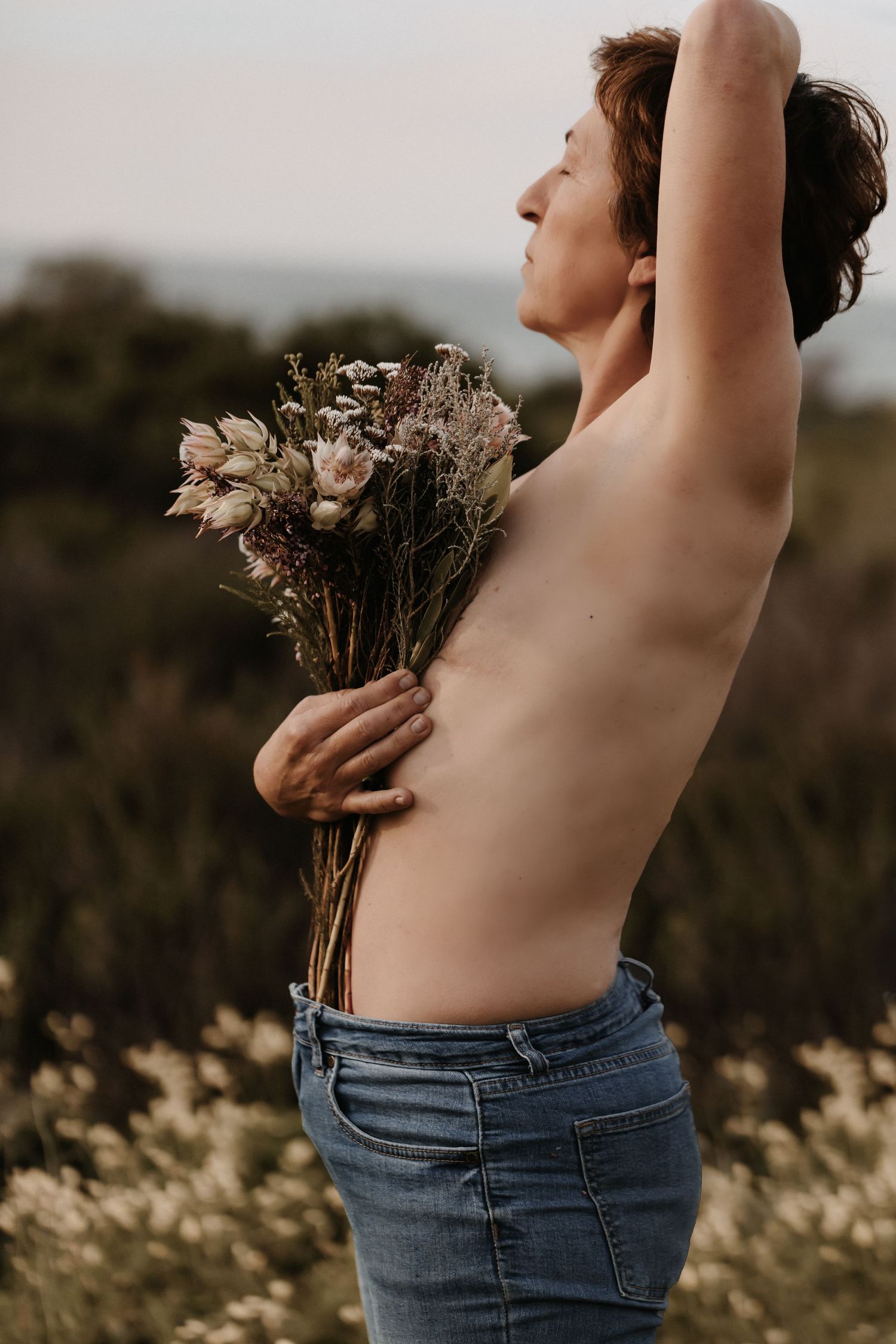 A person in blue jeans stretches their arms upwards while standing amongst blooming flowers at dusk.