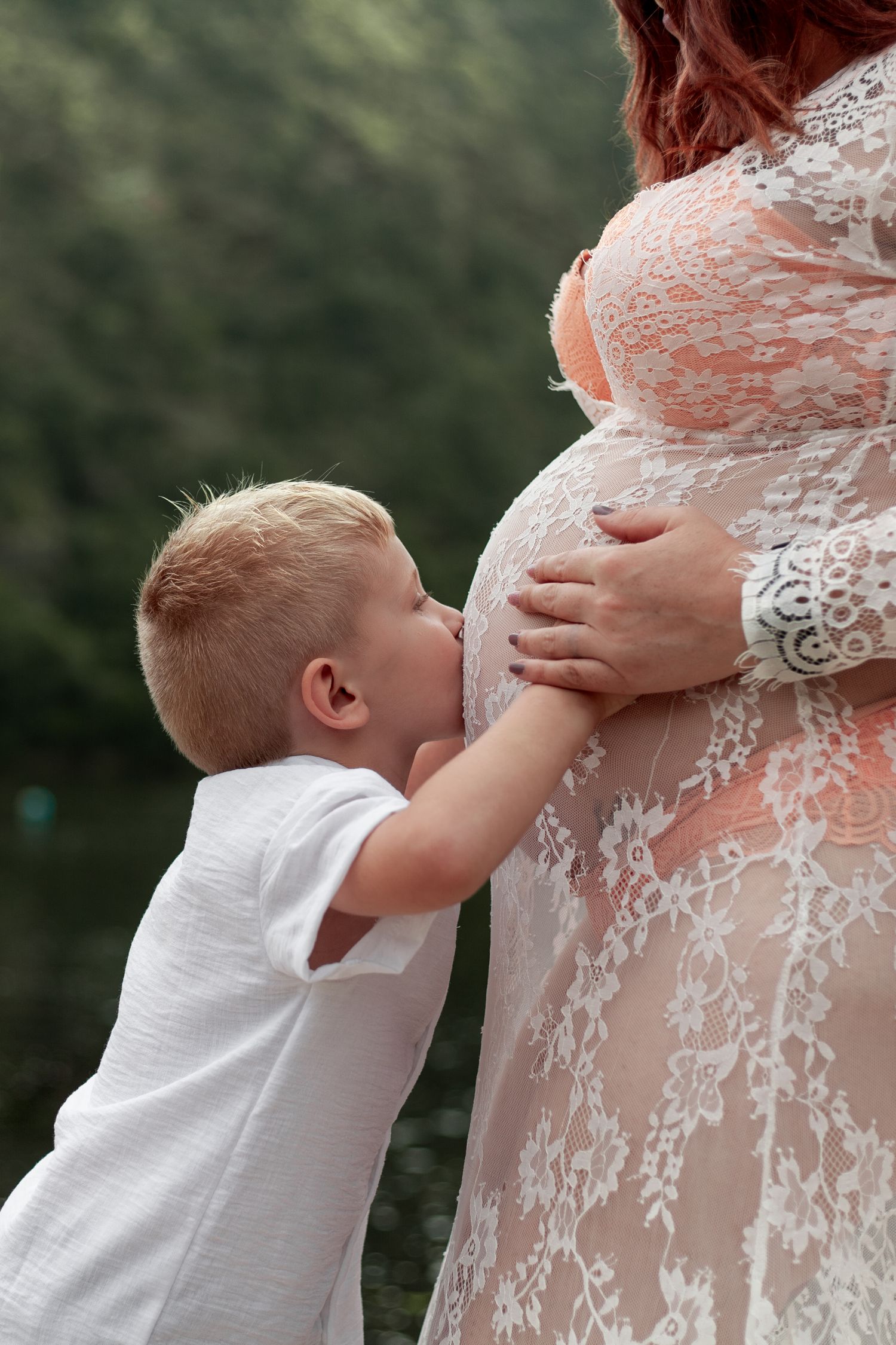 Young child in white shirt kisses pregnant woman's belly through delicate white lace dress in outdoor setting.