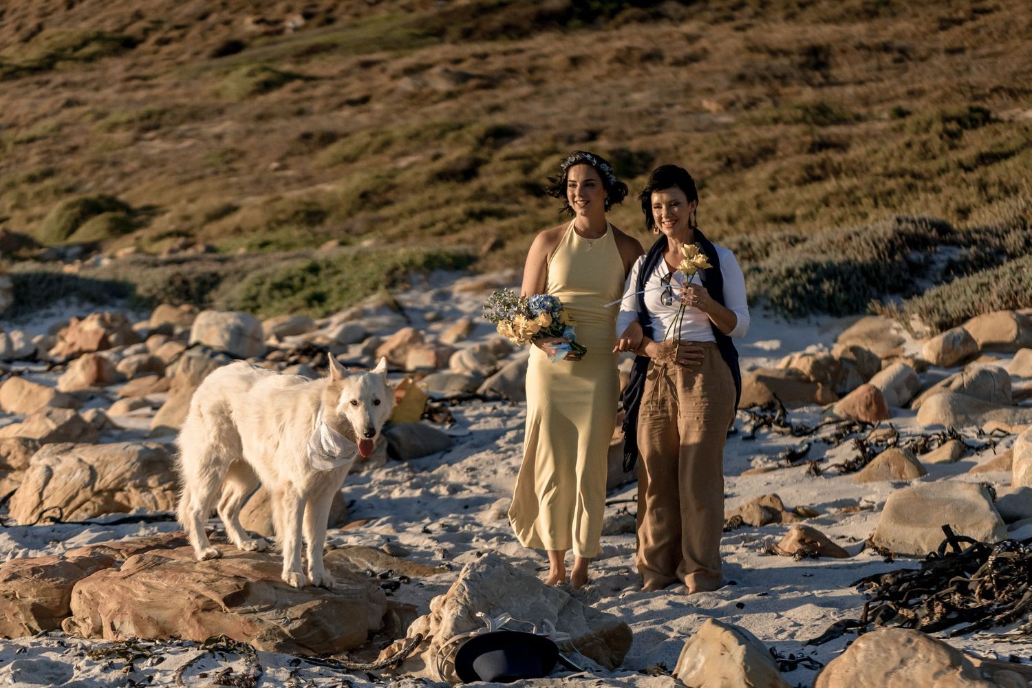 Two people stand with a large white dog on a rocky beach during sunset.