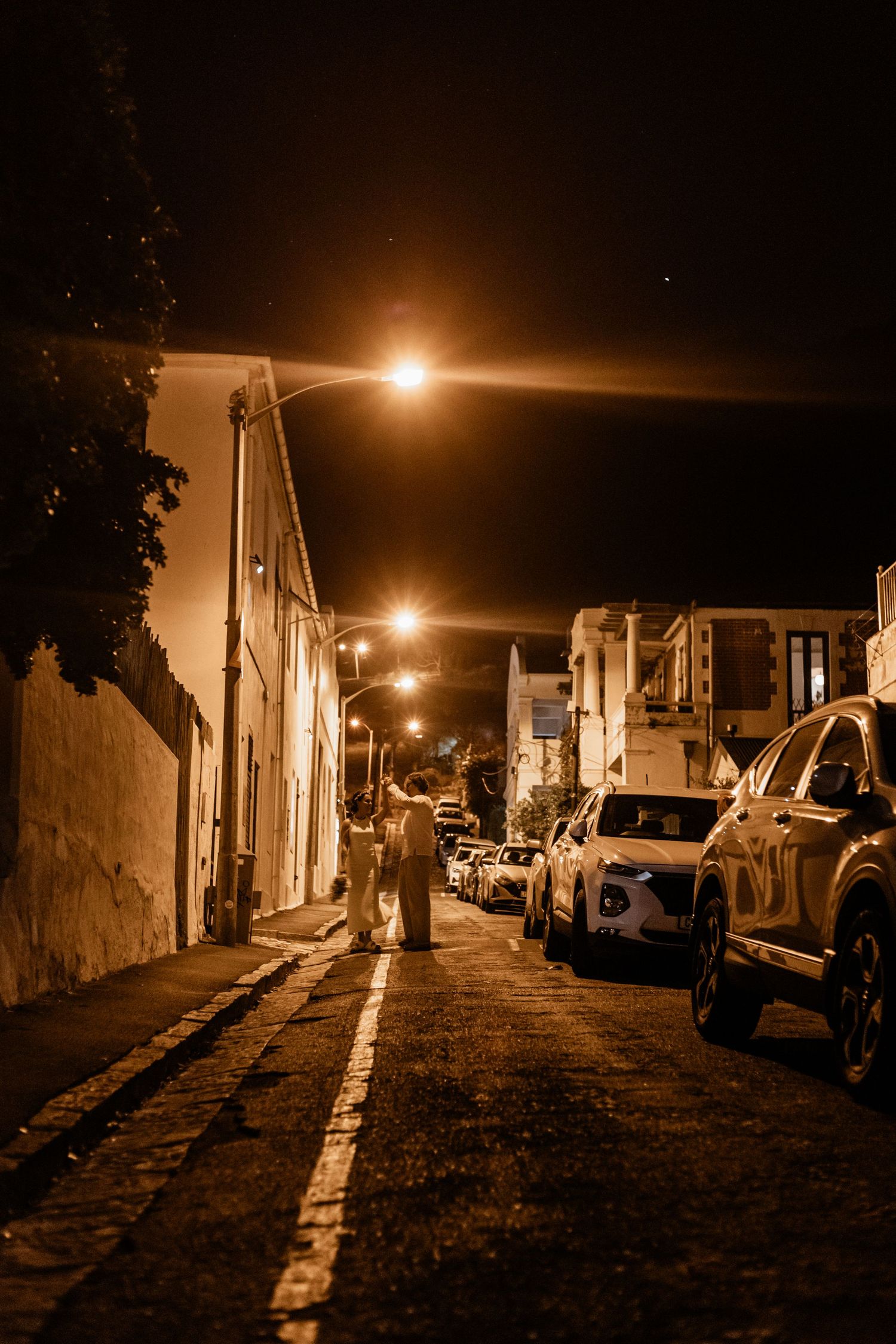 A person walks down a dimly lit street at night with cars parked along the curb under warm street lights.