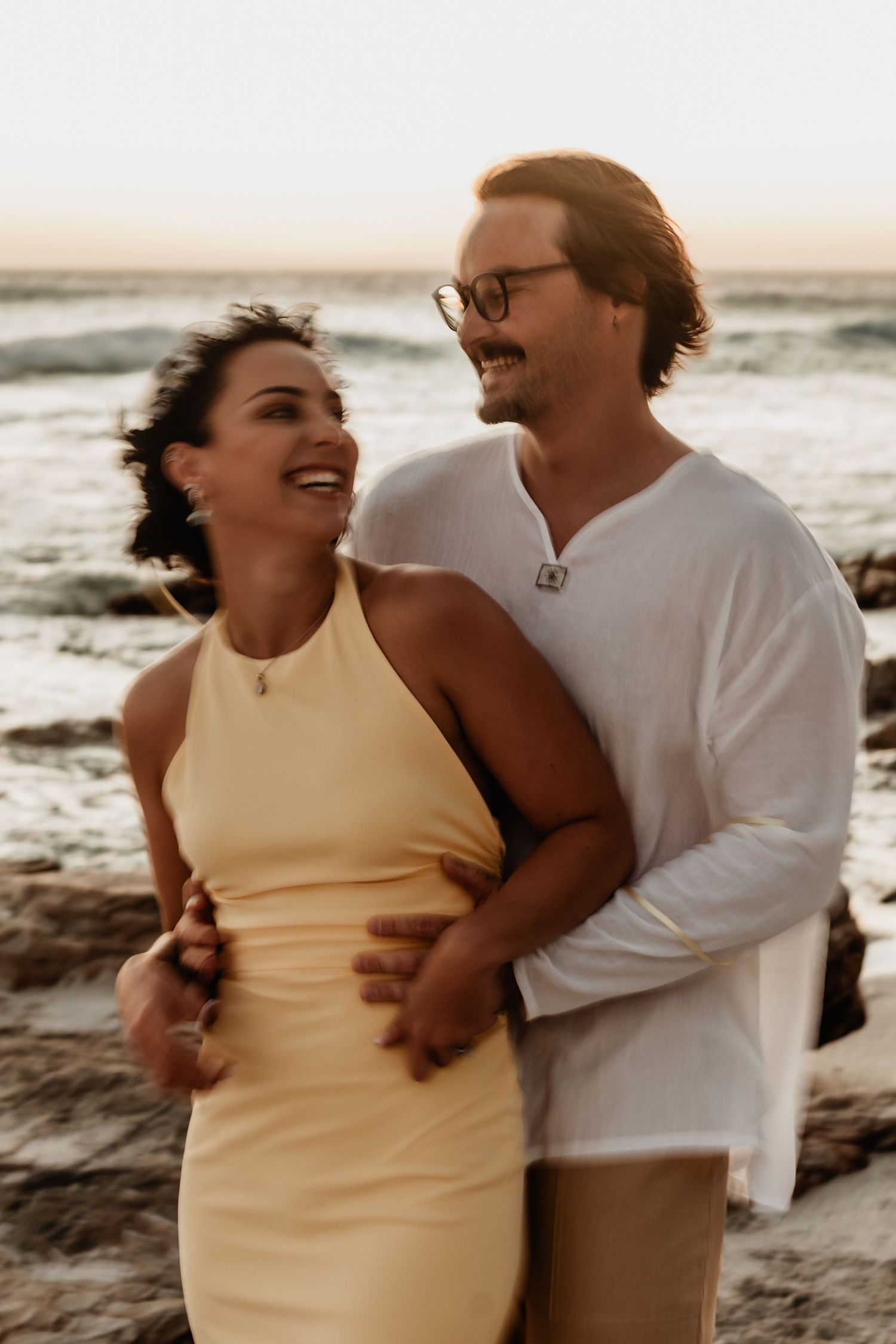 A happy couple embraces on the beach at sunset, sharing joyful laughter and tender moments.
