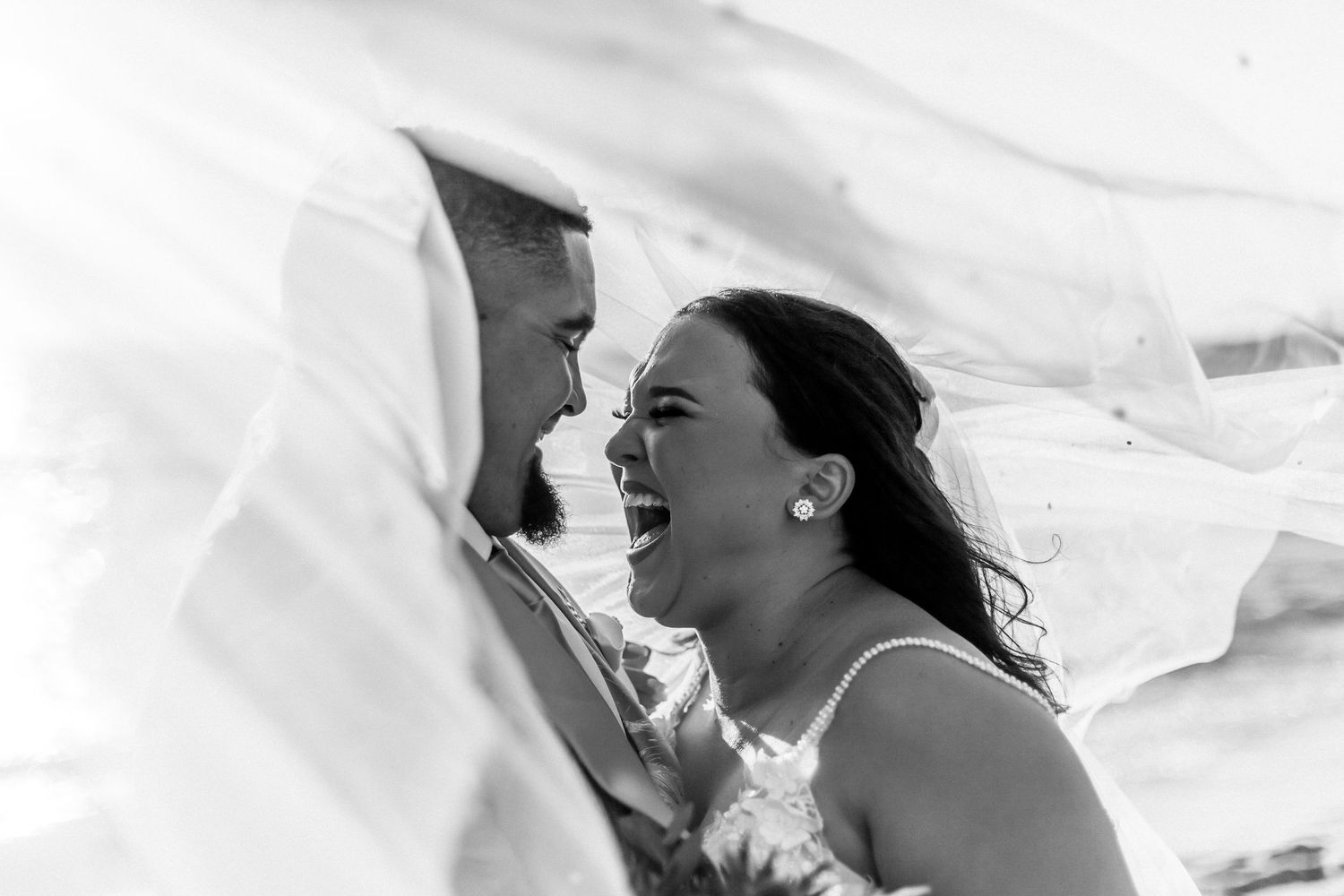 A black and white wedding photo capturing a joyous candid moment under flowing fabric veil.