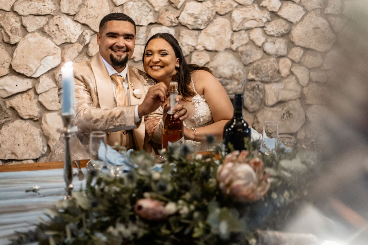 A happy couple laughs together at their wedding reception table decorated with pink floral arrangements and stone wall backdrop.