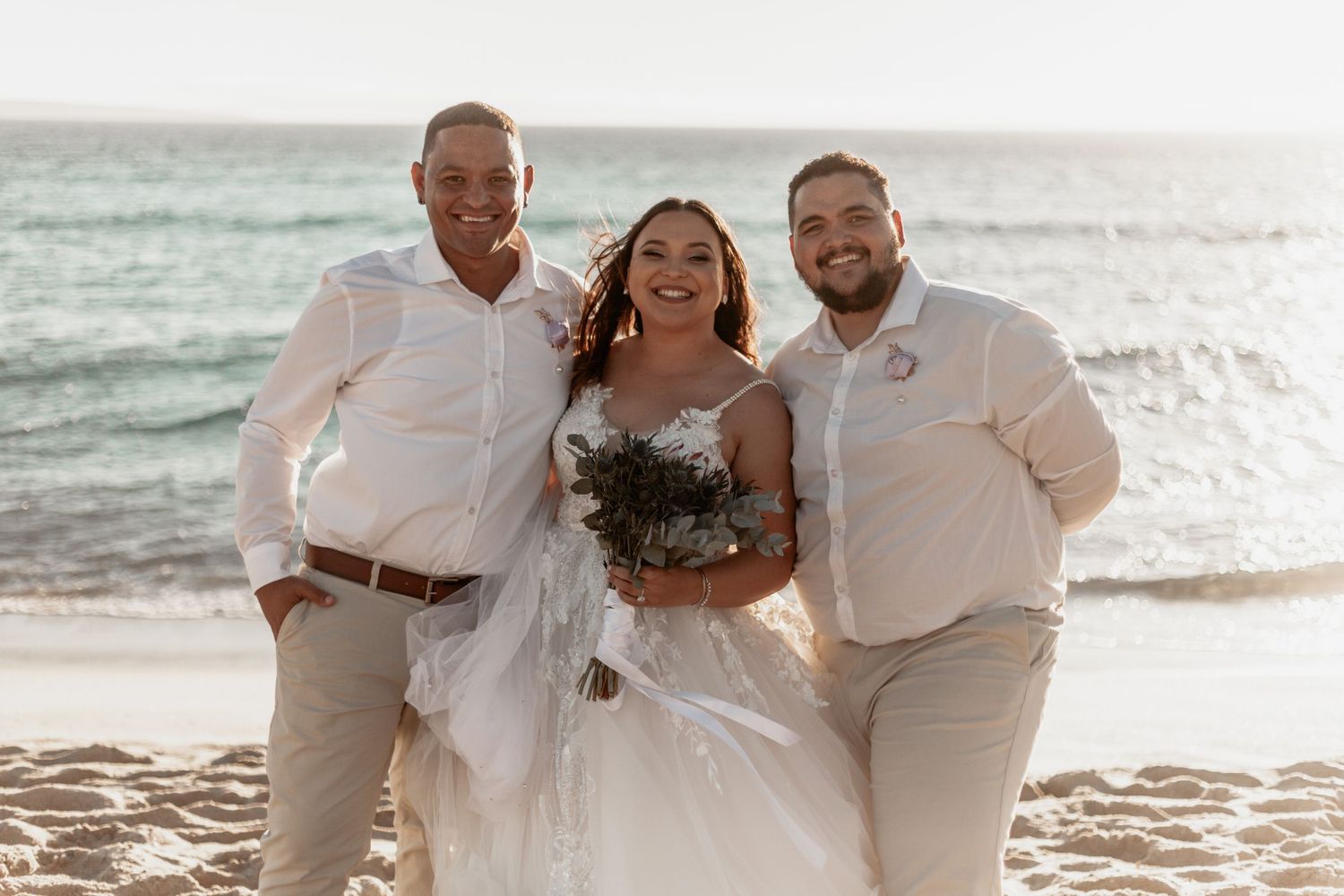 Beachside wedding celebration with three people in light colored outfits smiling on sandy shore at sunset.
