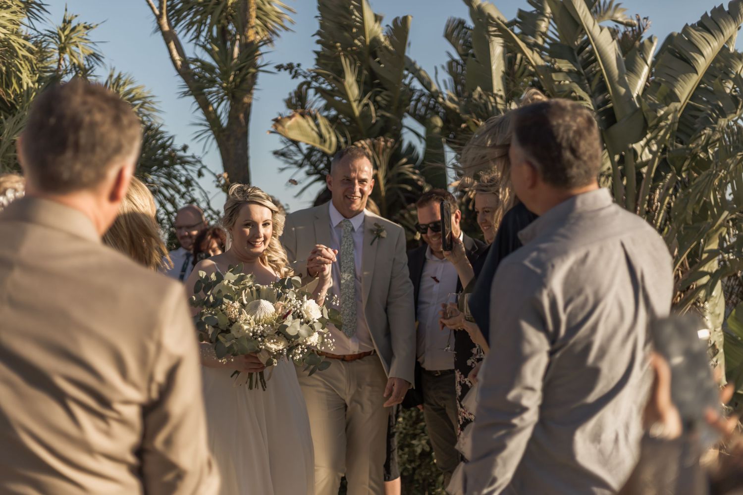A wedding ceremony takes place outdoors in a tropical setting with palm trees and banana plants in the background.