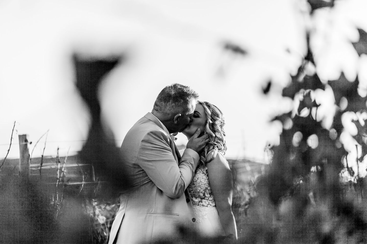 Black and white romantic photograph showing couple sharing an intimate kiss framed by soft bokeh foreground elements.