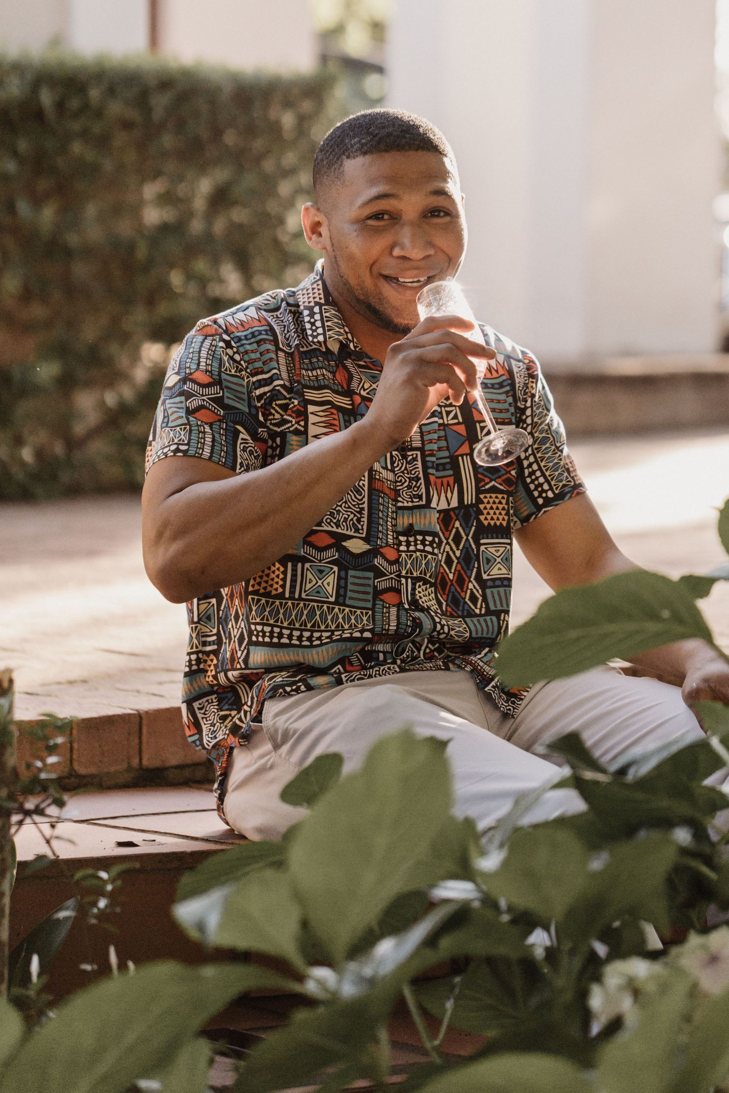 Someone wearing a geometric patterned short-sleeve shirt sits outdoors smiling while surrounded by green foliage.