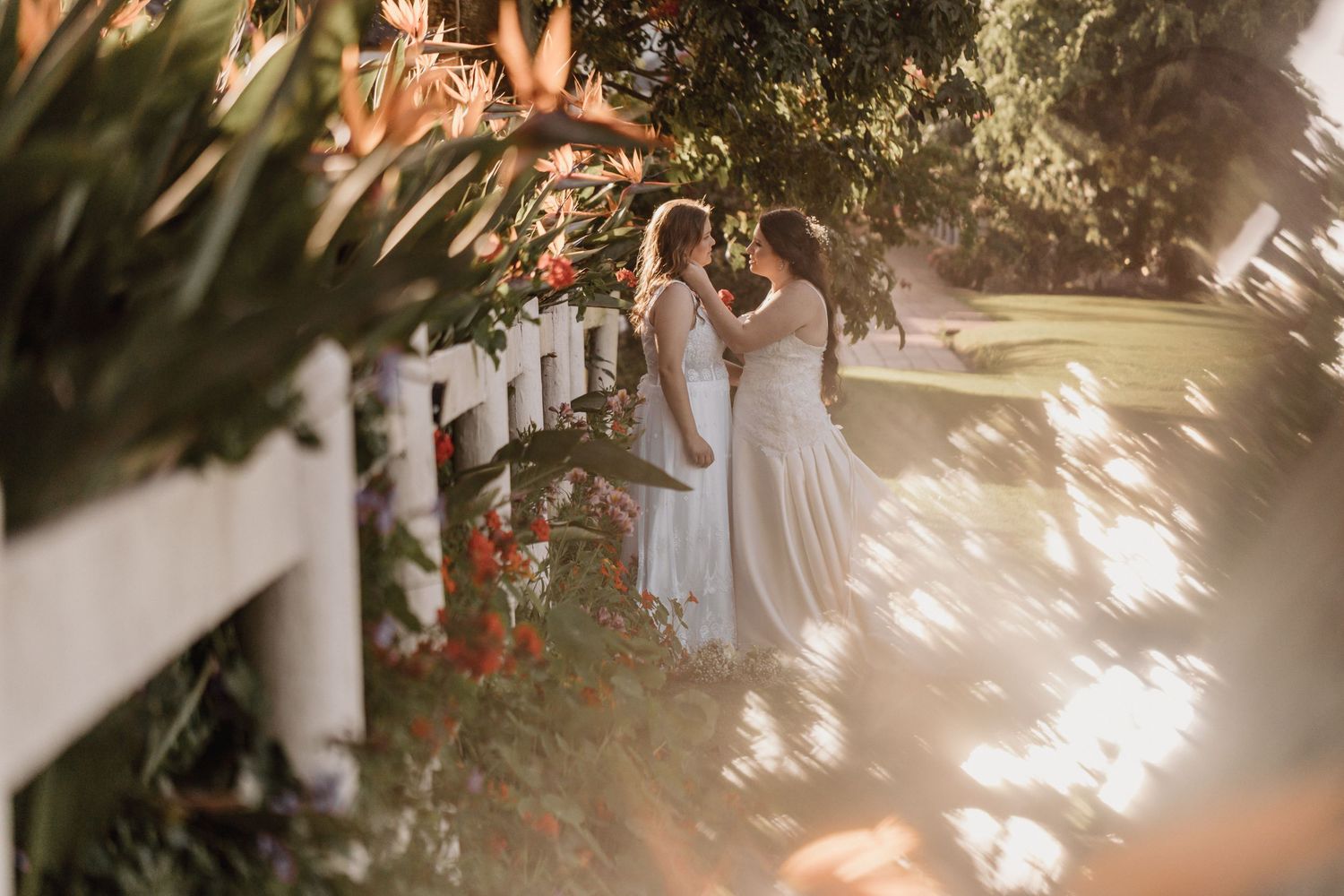 A sunlit garden wedding ceremony features white chairs and floral decorations under natural greenery and trees.