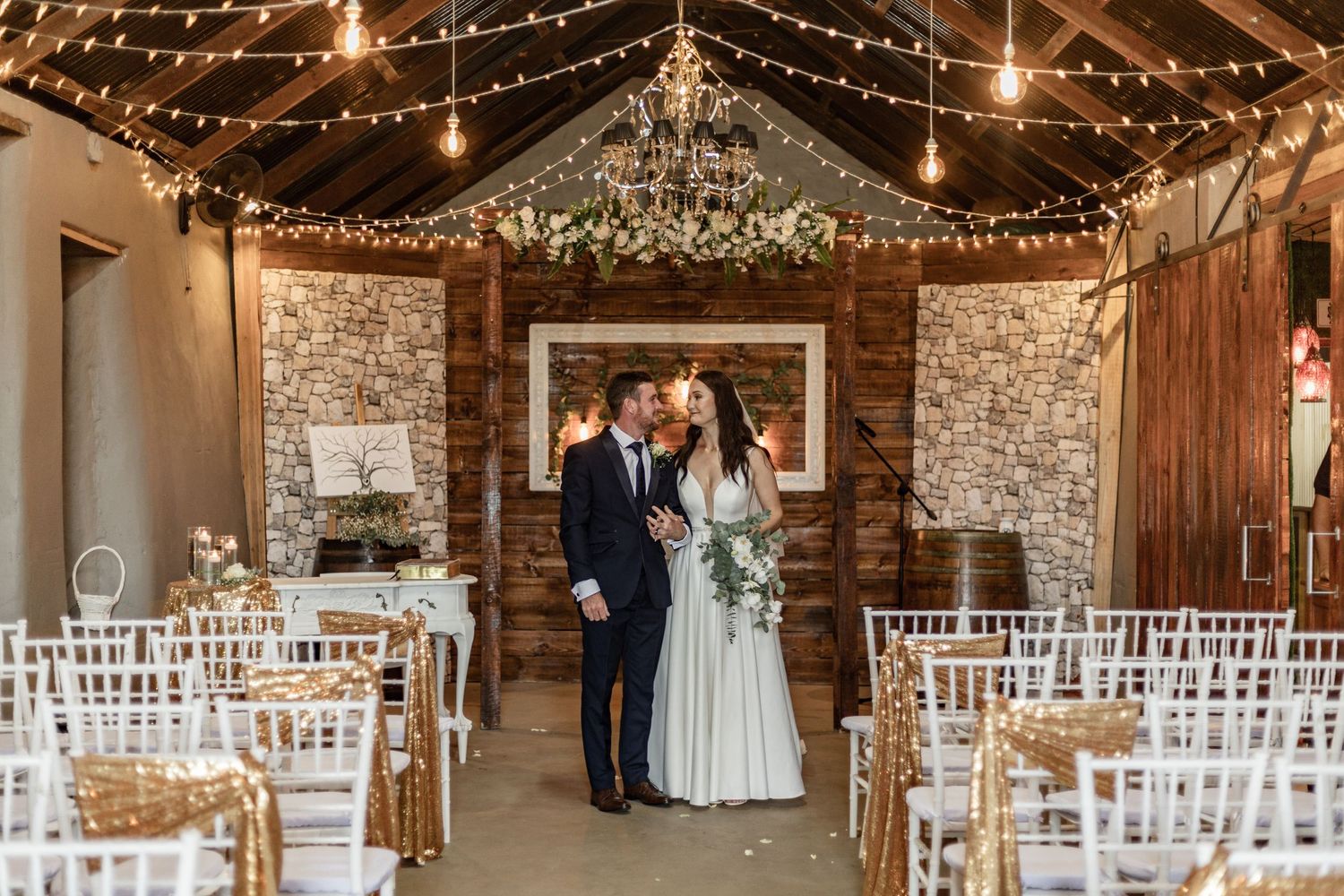 Rustic barn wedding venue decorated with string lights, crystal chandelier and white chairs in ceremony setup.