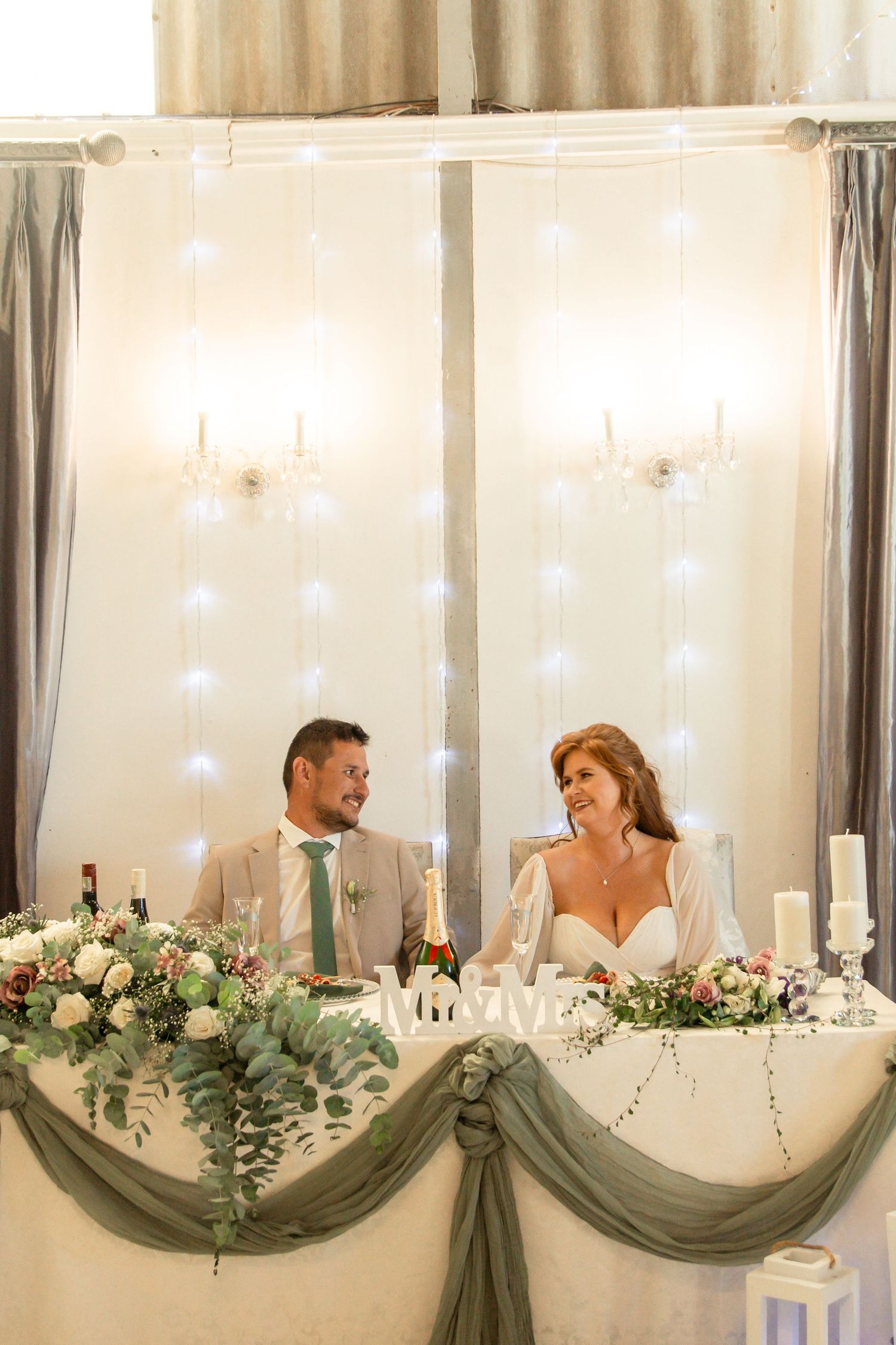 Bride and groom smiling at sweetheart table decorated with sage green draping and floral garlands at wedding reception.