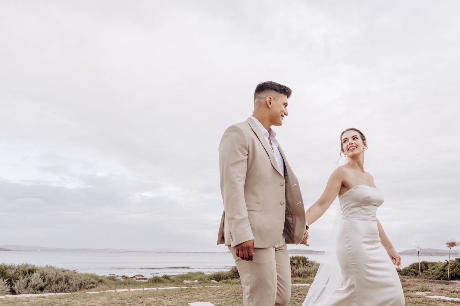 A smiling bride and groom hold hands walking outdoors on their wedding day with an overcast coastal backdrop.