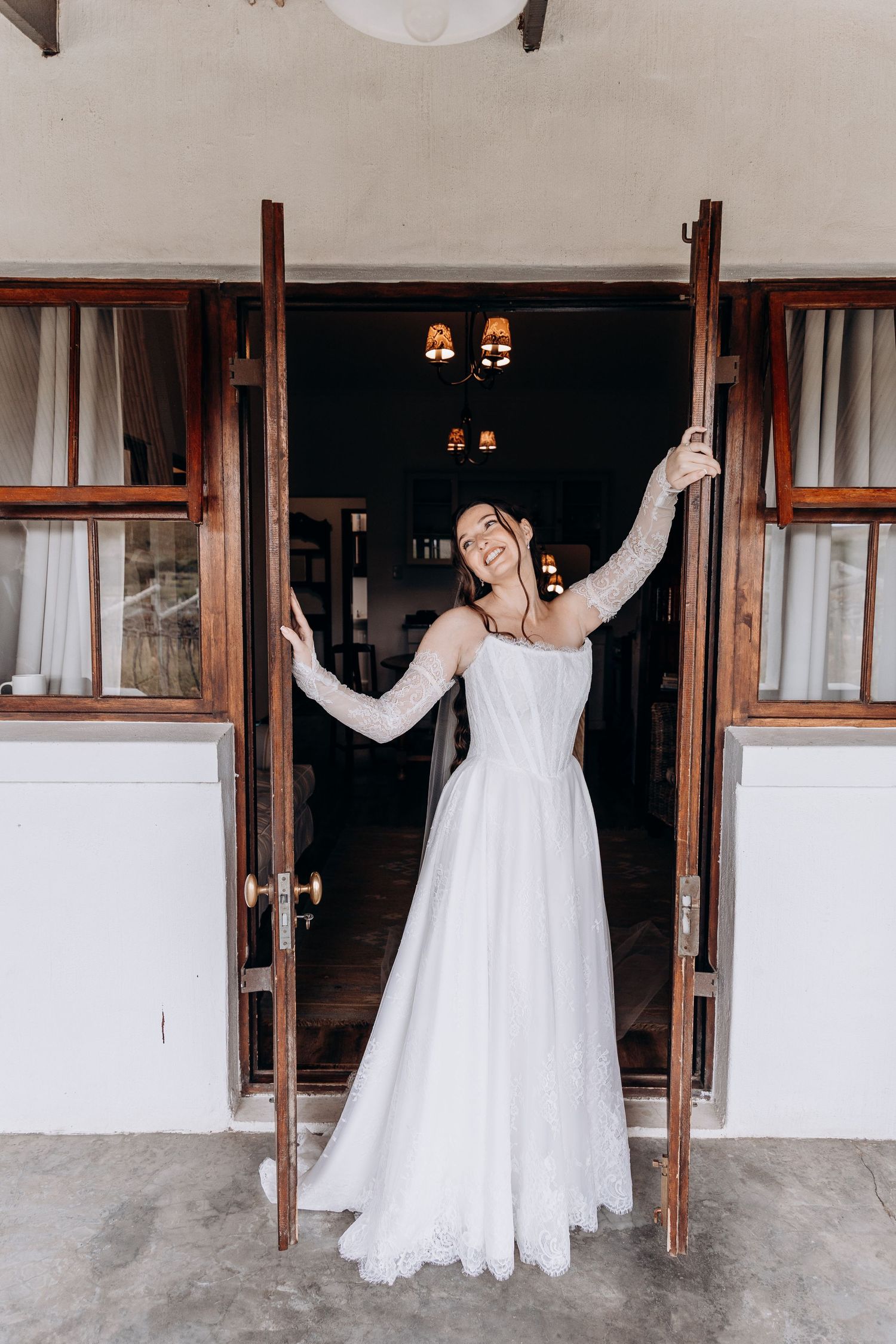 Smiling bride in white lace gown with long sleeves poses in open wooden double doors of a rustic venue.